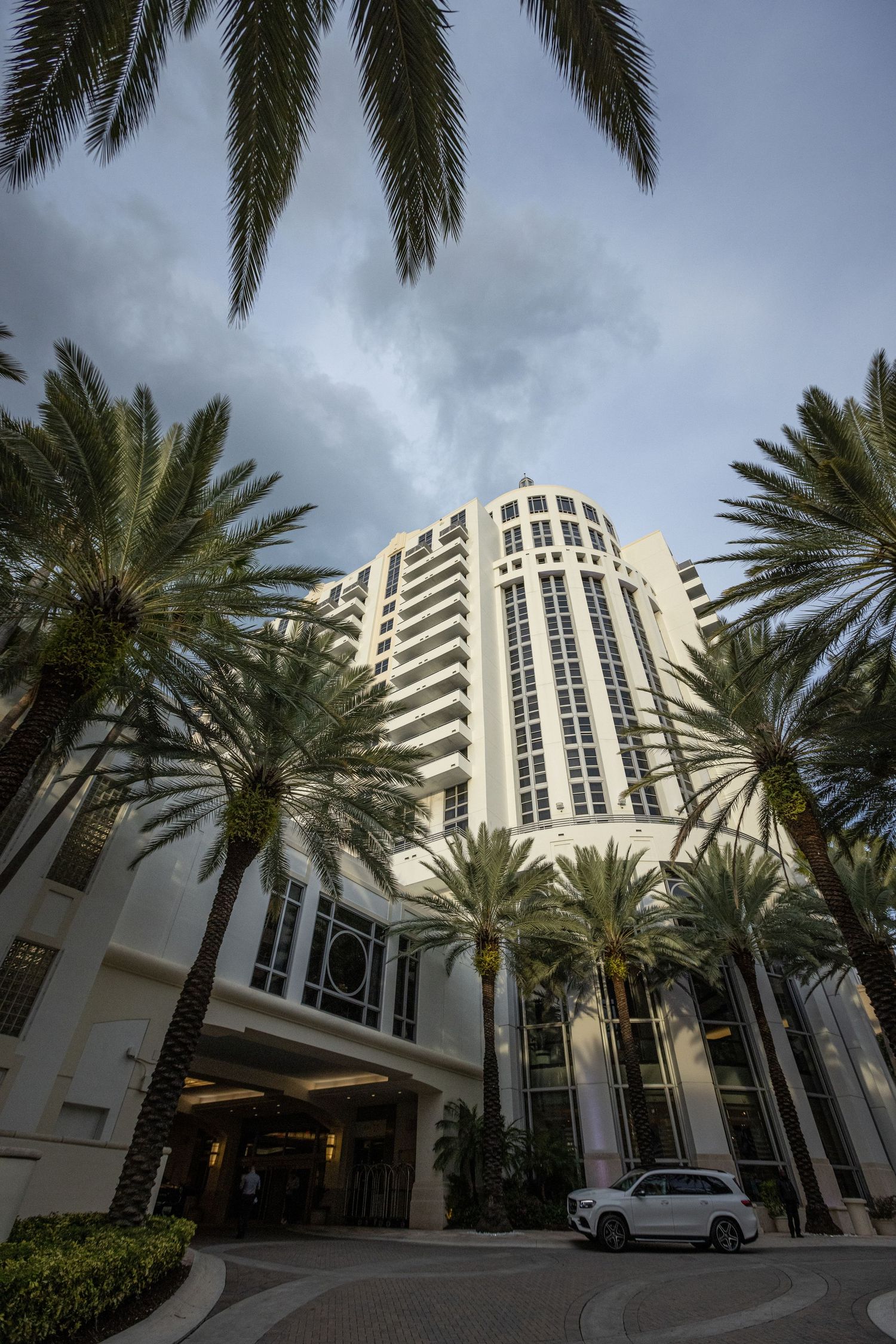 Modern white commercial building with palm trees and curved driveway entrance under dramatic cloudy sky in Miami.