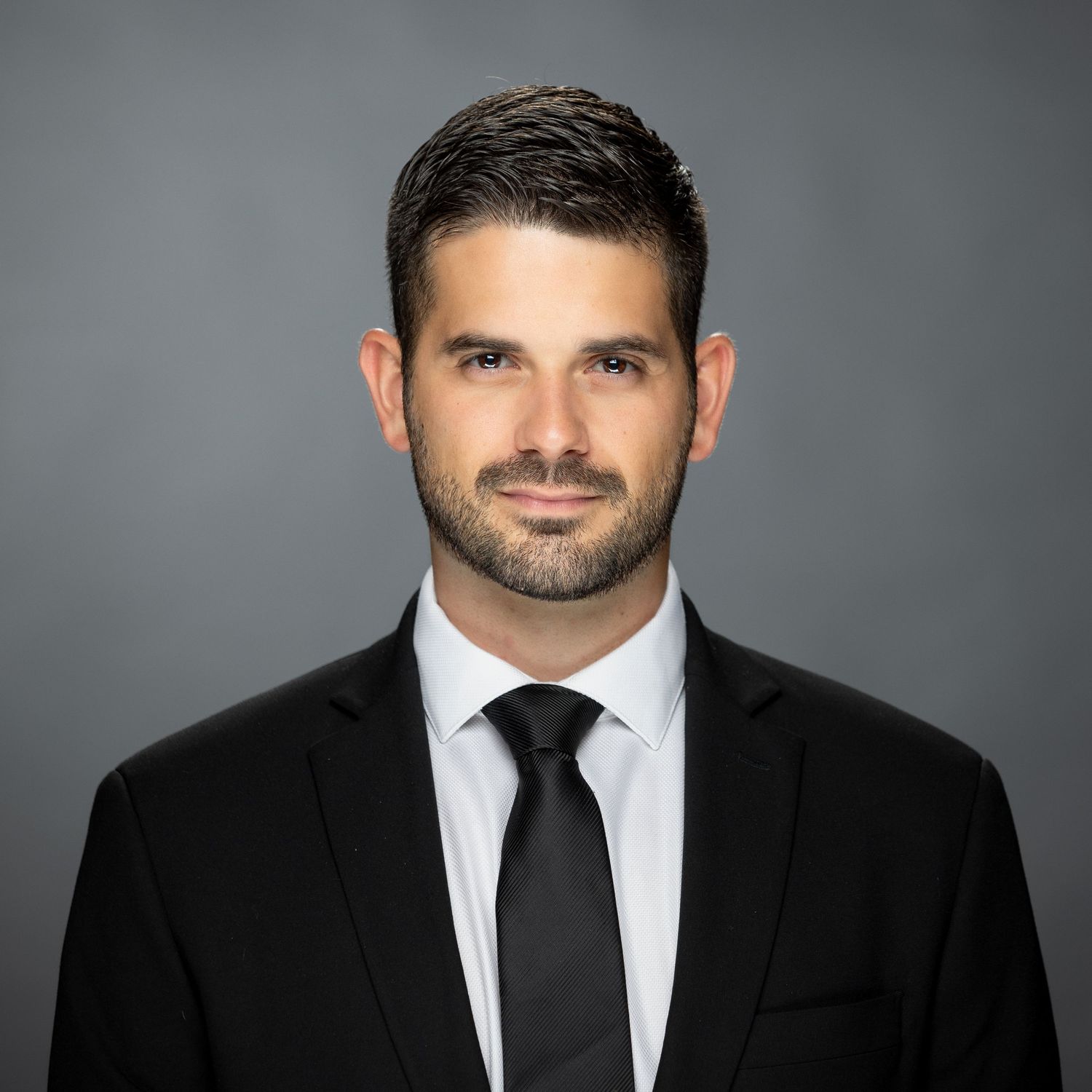 Professional business portrait featuring someone wearing a black suit and tie against a gray backdrop.