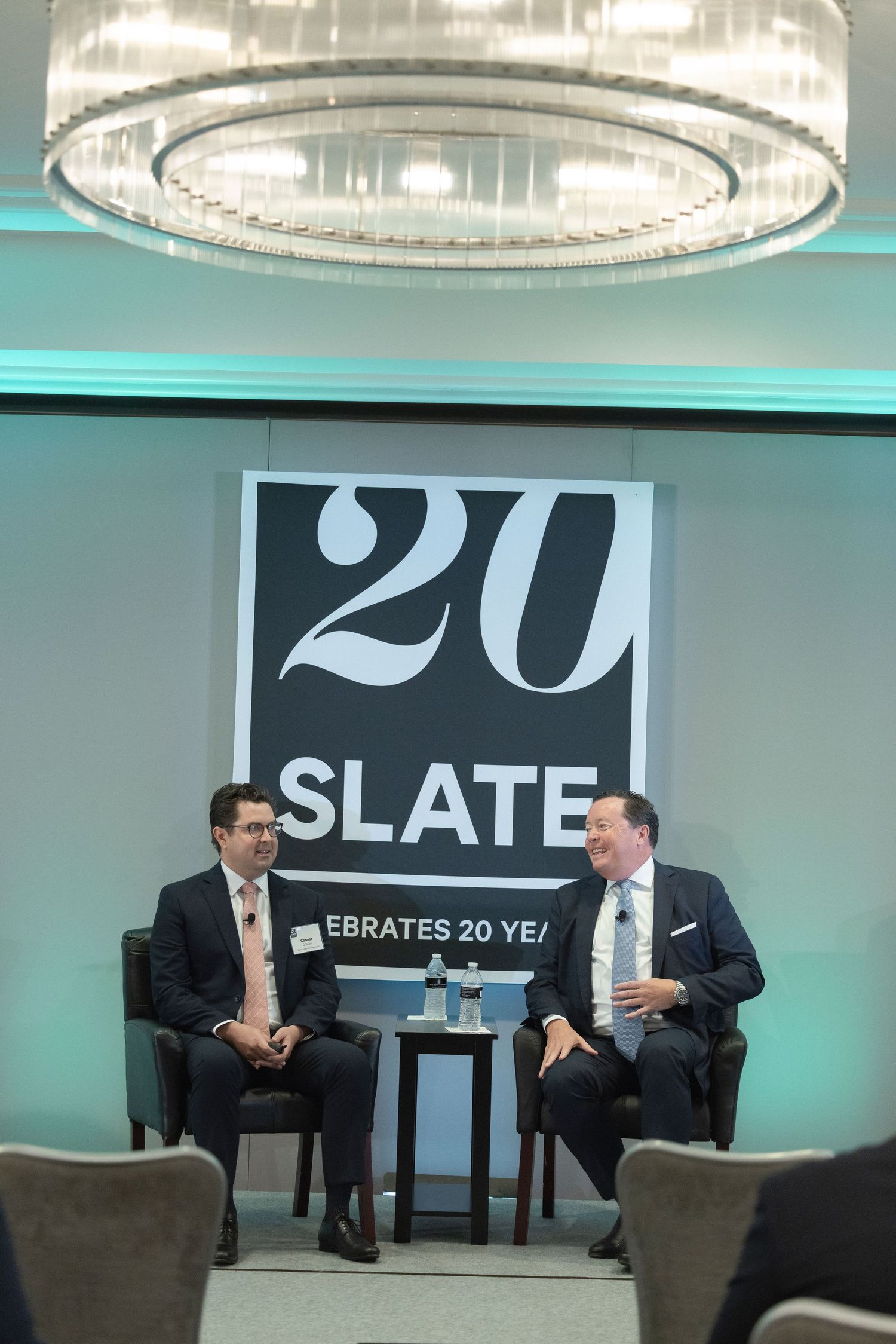 Two people sit in armchairs on stage during what appears to be a professional speaking event with Slate branding.