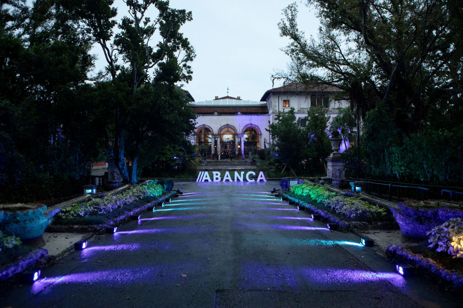 Long lighted walkway with illuminated benches and purple LED ground lighting leading to a garden space at dusk.