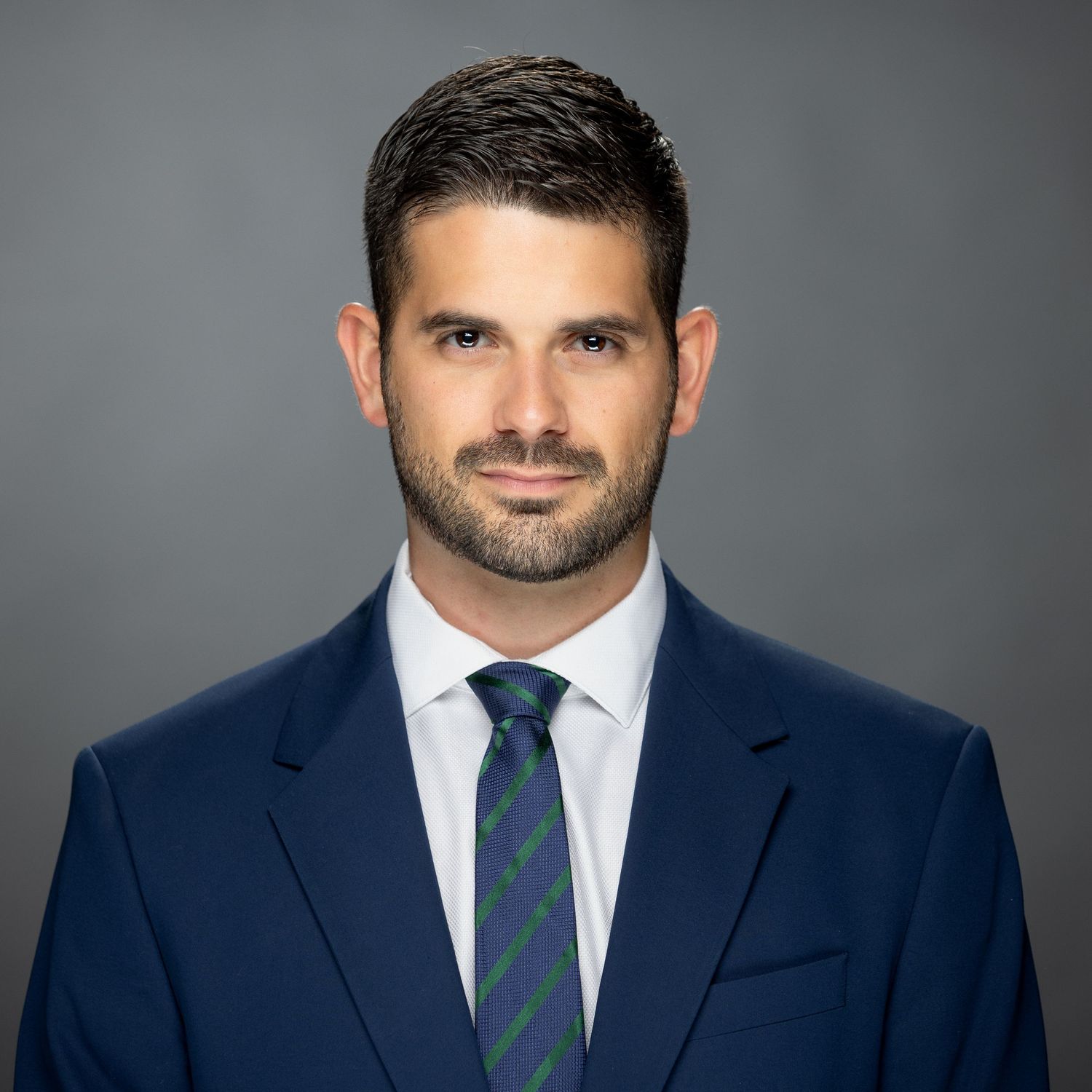 Professional headshot of someone in a navy suit with a green and navy striped tie against a gray background.