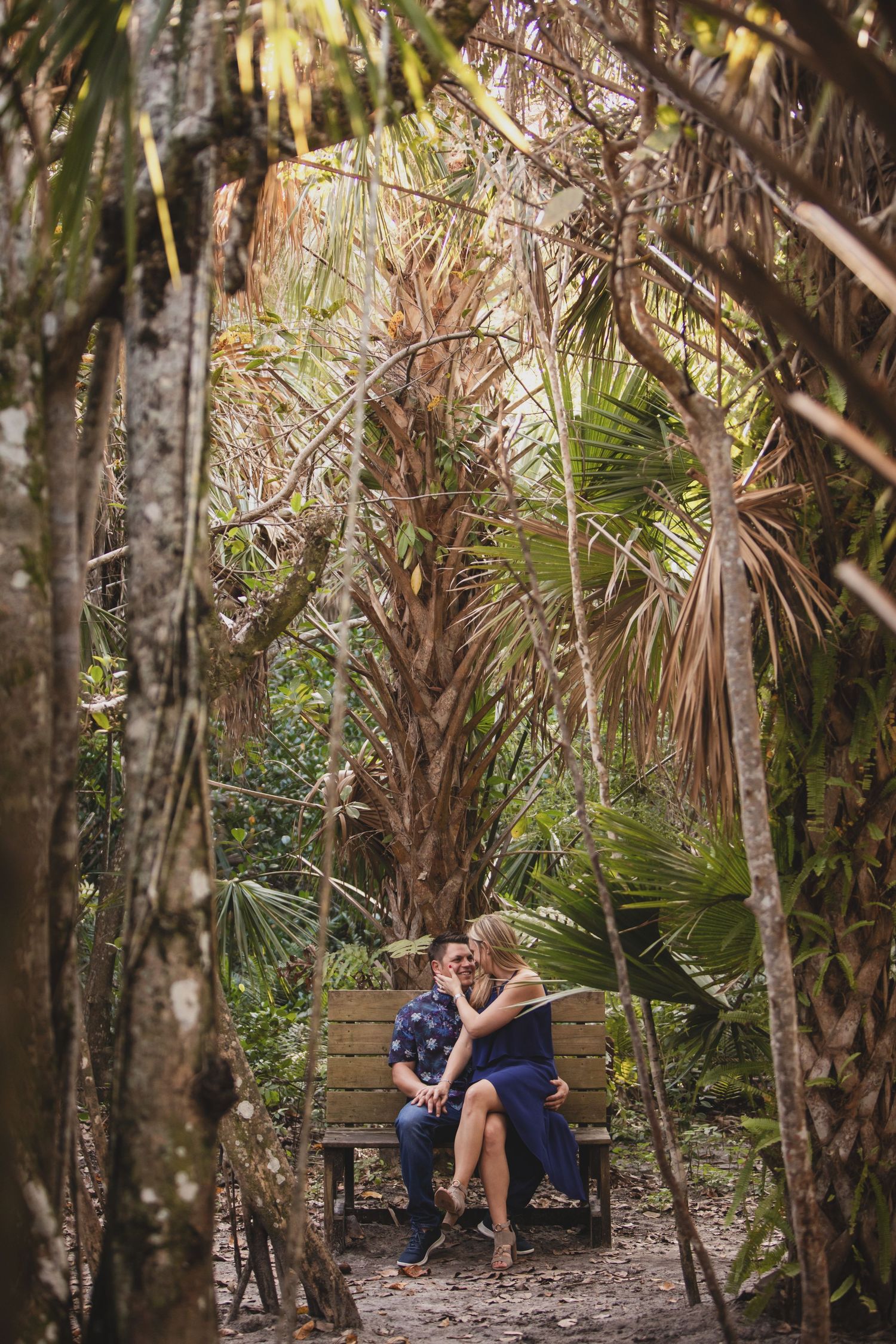 A couple shares an intimate moment on a wooden bench surrounded by tropical palm trees and foliage.