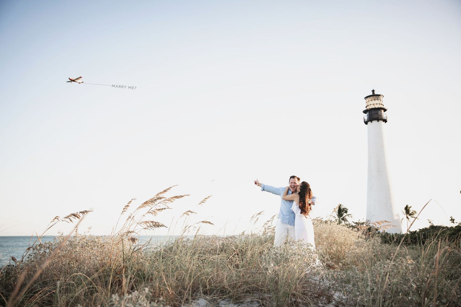 Lighthouse and seagrass on a coastal beach during daytime with birds flying overhead.