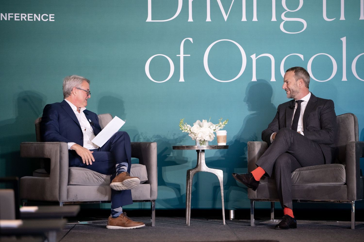 Two people engage in a business discussion while seated in gray armchairs on a stage with teal backdrop lighting.
