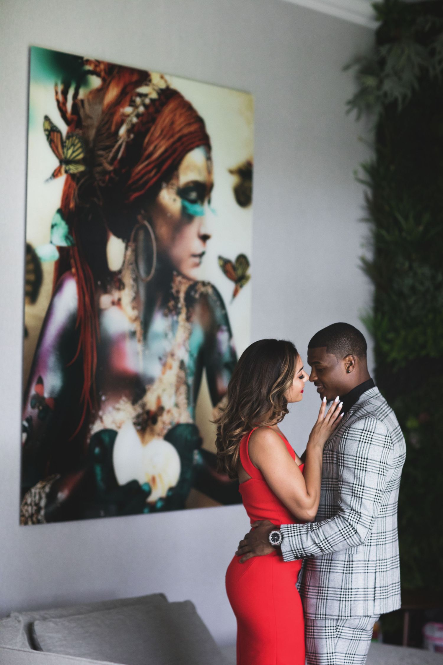 A couple poses in front of artistic portrait wall art, with person in red dress and person in checkered shirt embracing.