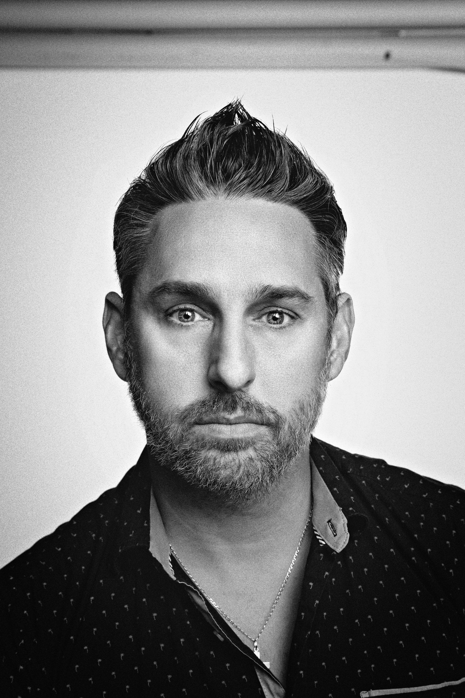 Black and white portrait of a bearded man wearing a dark polka dot shirt and necklace against a plain background.