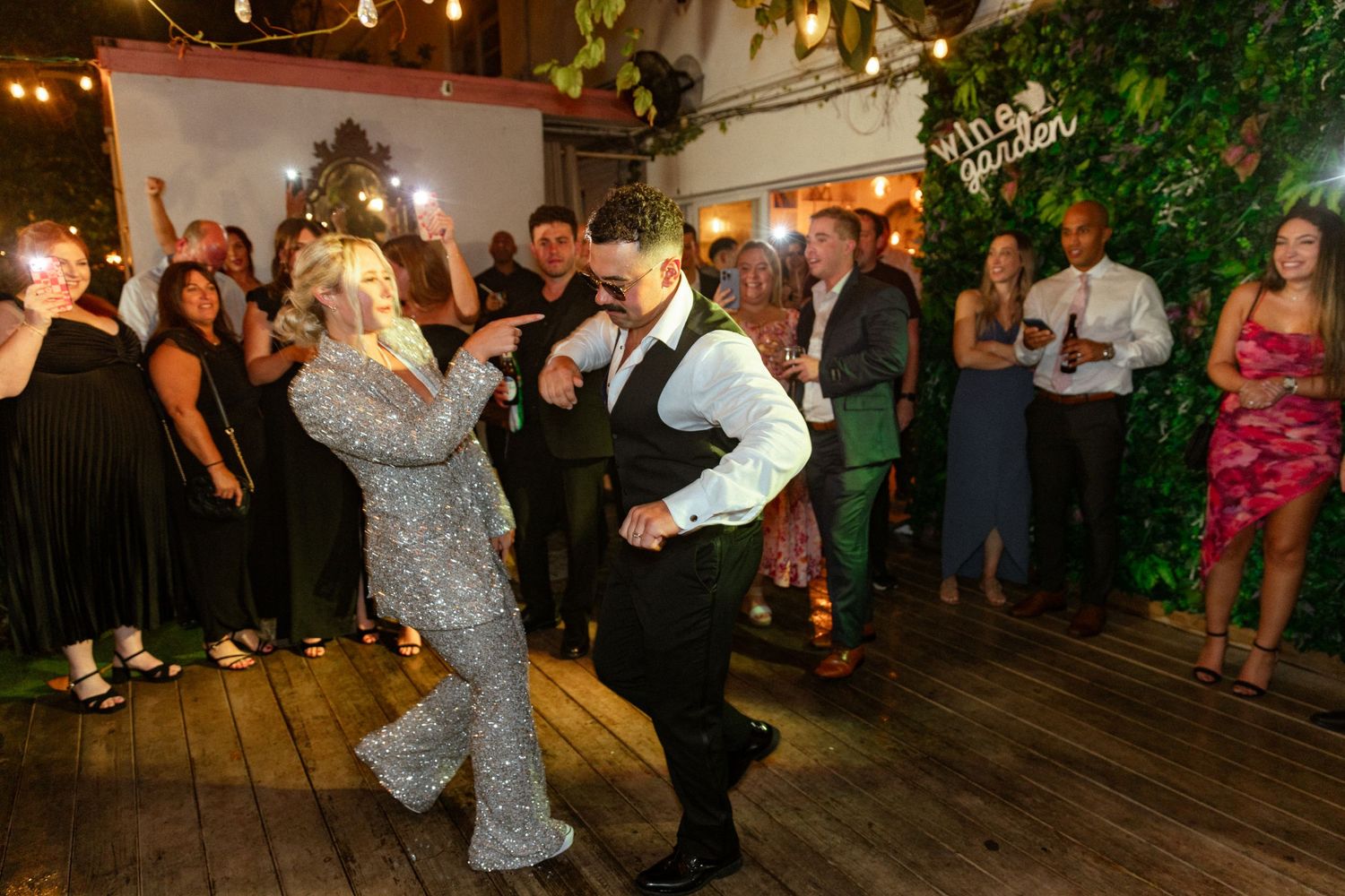Wedding guests dancing and celebrating on a festive dance floor with decorative lighting.