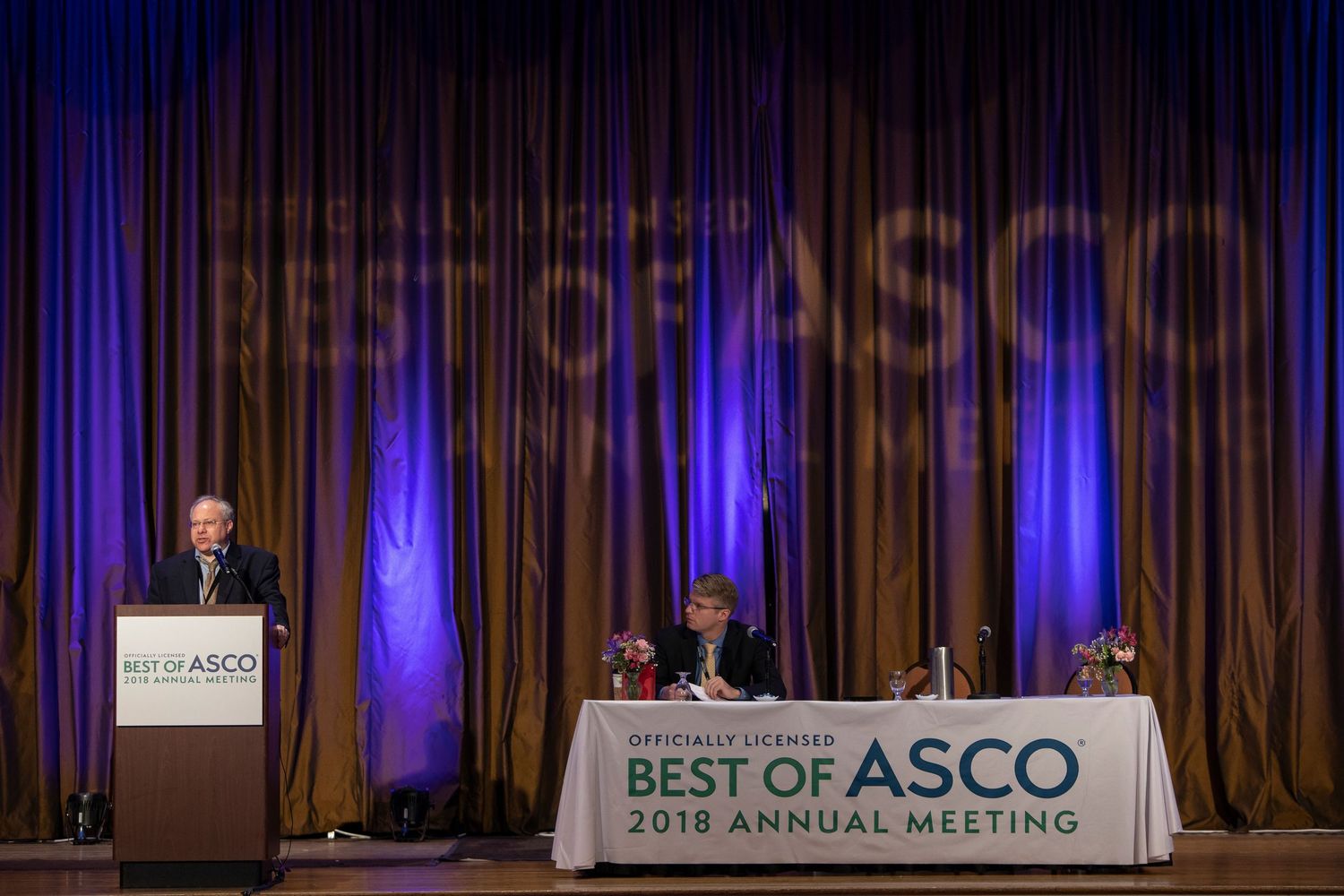 Speakers present at a medical conference podium with 'Best of ASCO' banner and blue-lit backdrop.