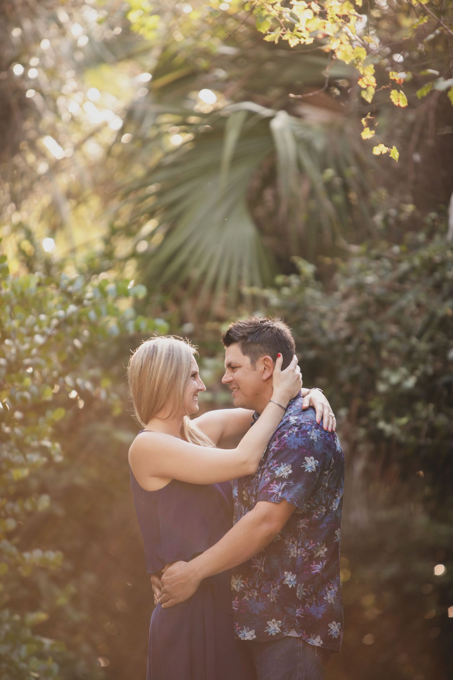A romantic couple embraces outdoors during golden hour beneath sunlit trees and palms.