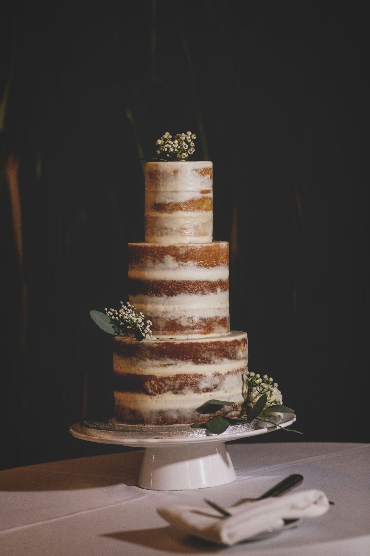 Rustic naked wedding cake with three tiers decorated with small white flowers on white tablecloth.