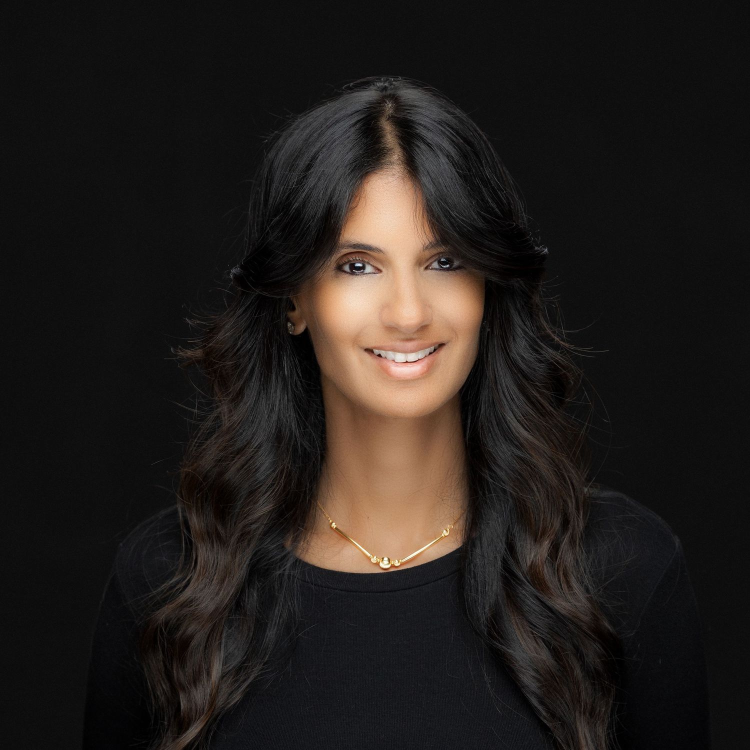 Woman with long dark hair and black top against dark background smiling warmly at camera in professional headshot.