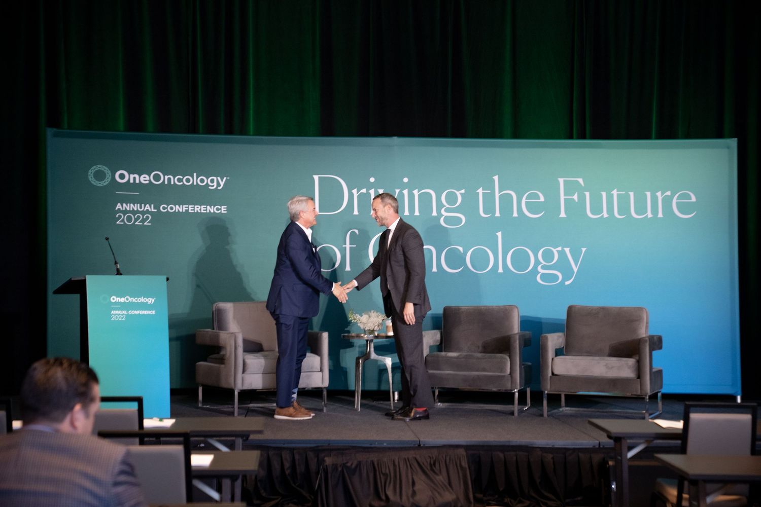 Business conference stage setup with modern teal backdrop showing 'Driving the Future' text and comfortable seating arrangement.