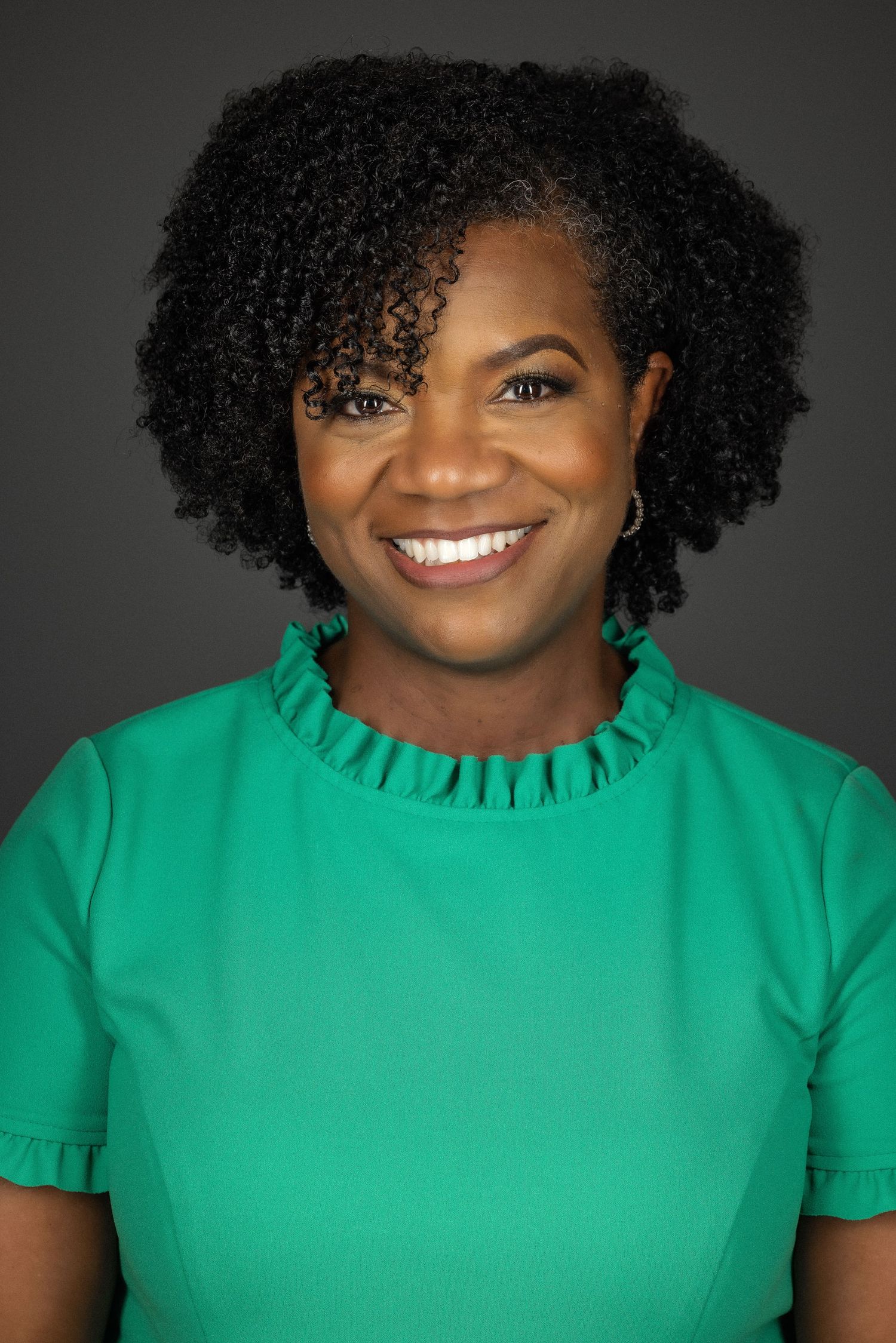 Portrait against dark background showing bright emerald green ruffle neck top and natural curly hairstyle with joyful expression.