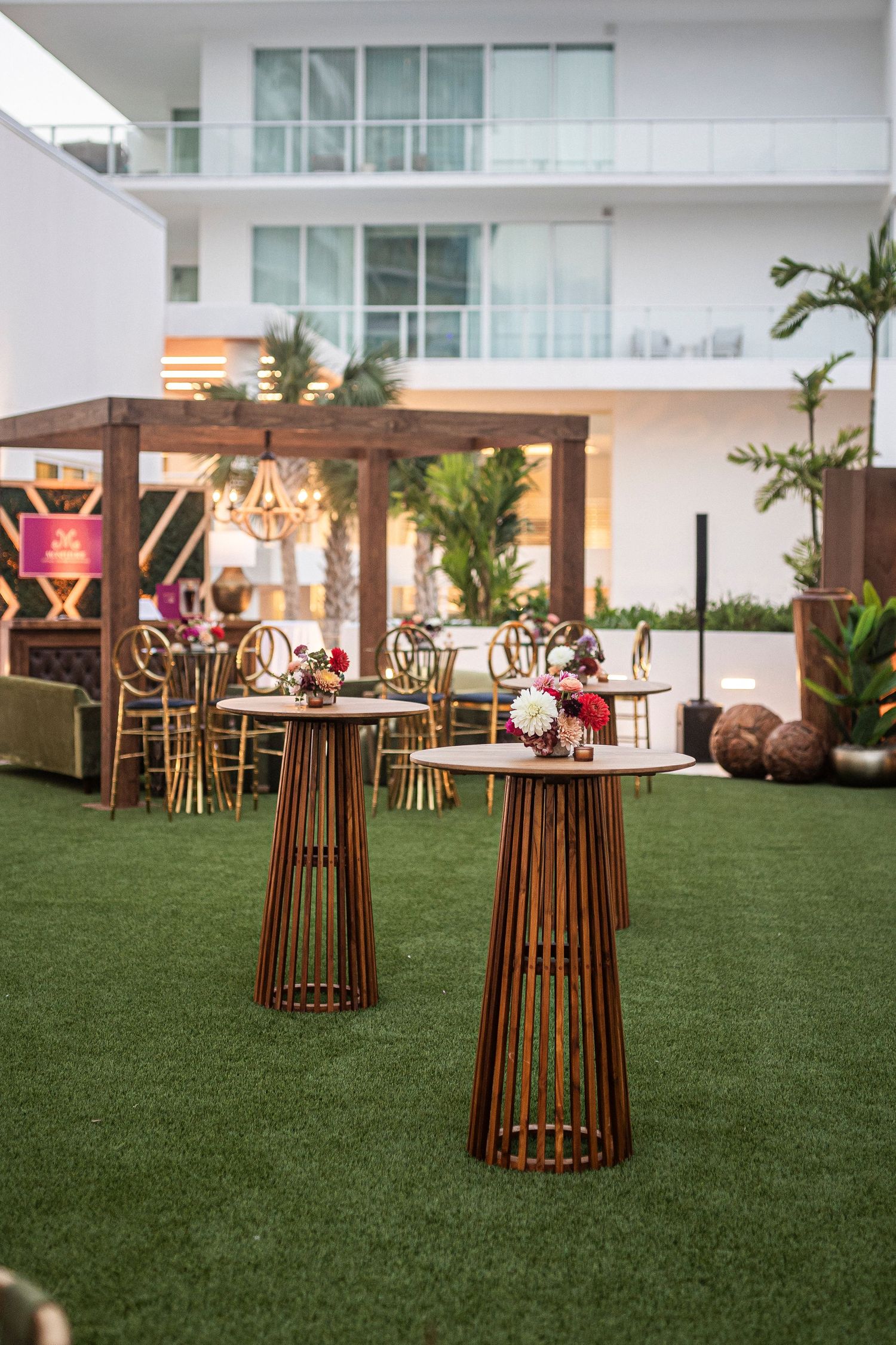 Modern outdoor event setup with copper cocktail tables and chandelier on artificial grass terrace at luxury venue.