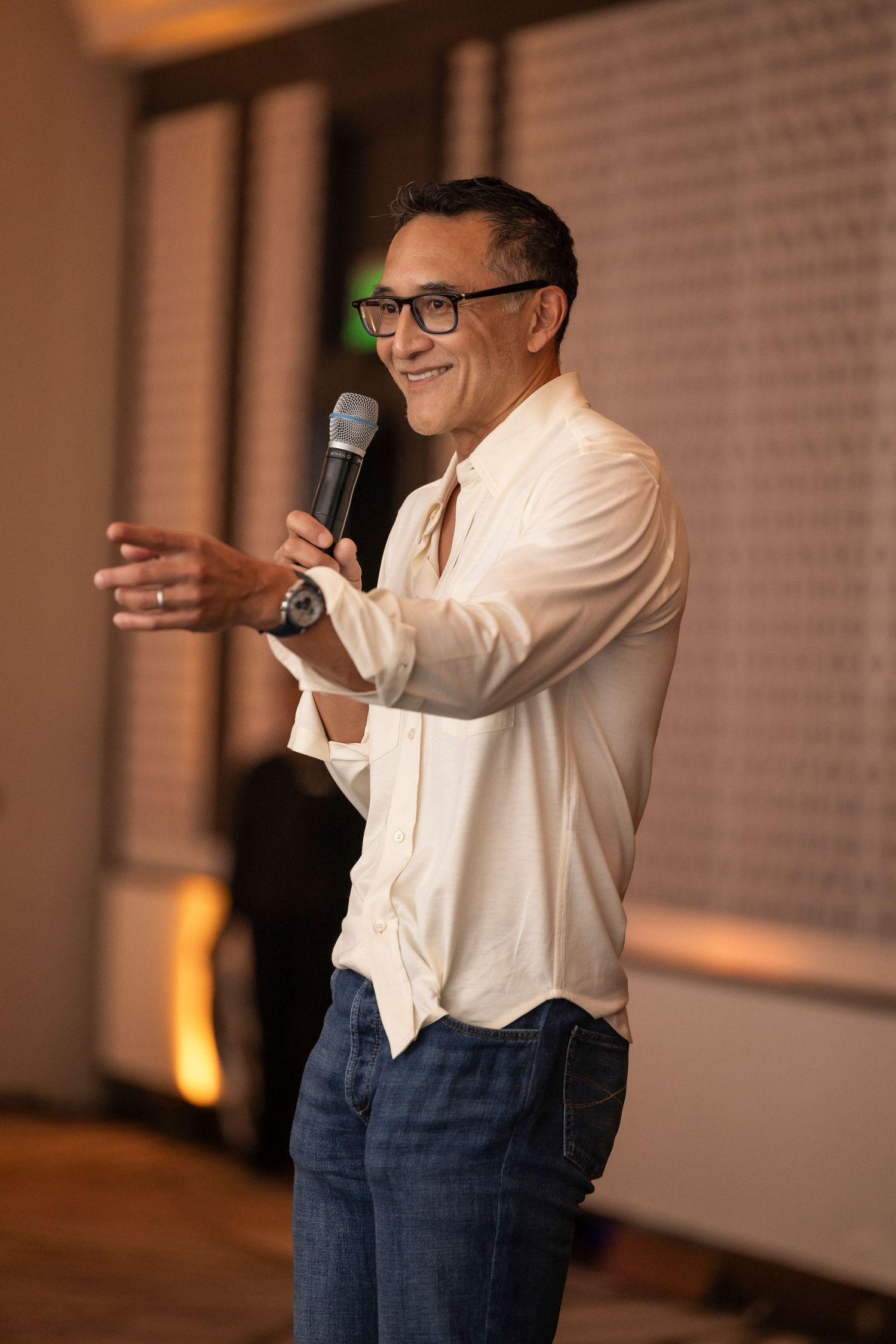 A smiling speaker wearing glasses and a white button-down shirt gestures while holding a microphone on stage.