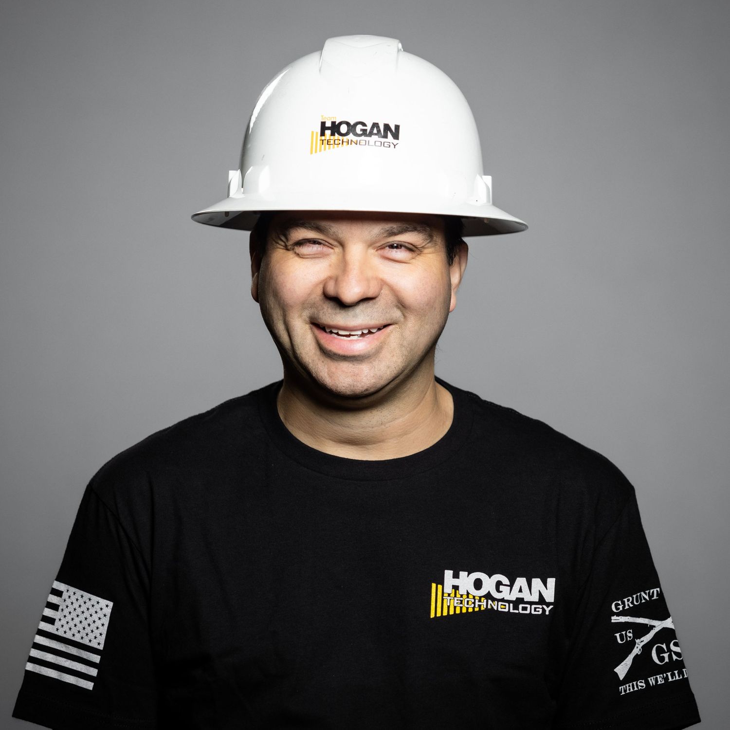 Construction worker wearing Hogan branded hardhat and t-shirt smiling against gray background.