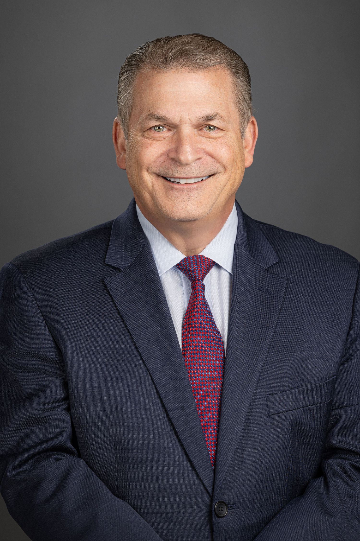 Professional headshot of a person in a dark navy suit and red tie smiling against a gray background.