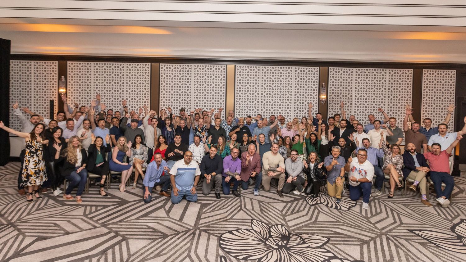 A large panoramic group photo of conference attendees sitting and standing in rows in a modern hotel ballroom.