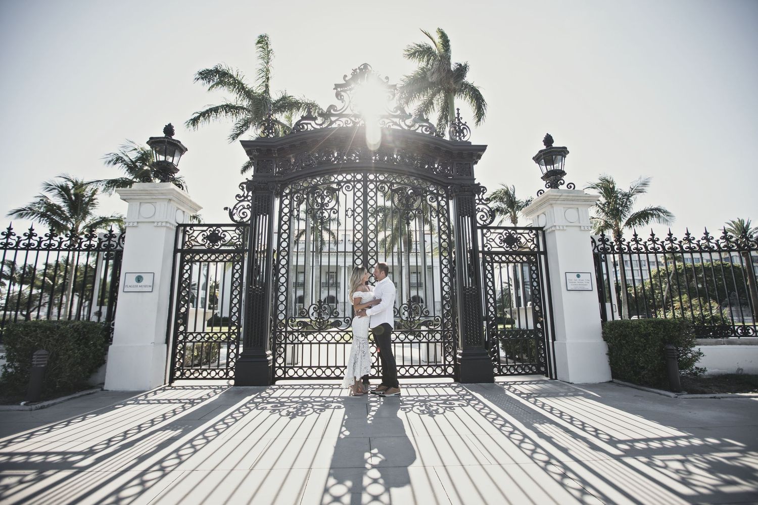 Ornate wrought iron gates cast dramatic shadows on a white driveway lined with palm trees.