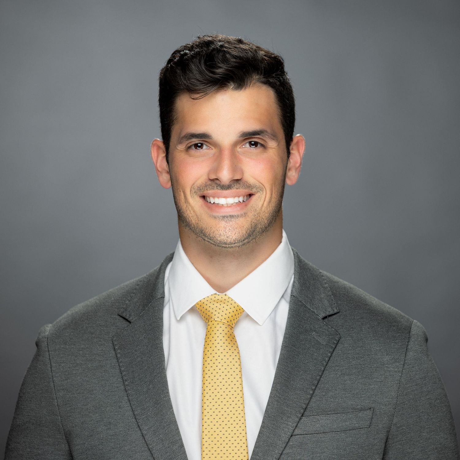 Professional headshot of someone wearing a gray suit and bright yellow tie against a neutral background.