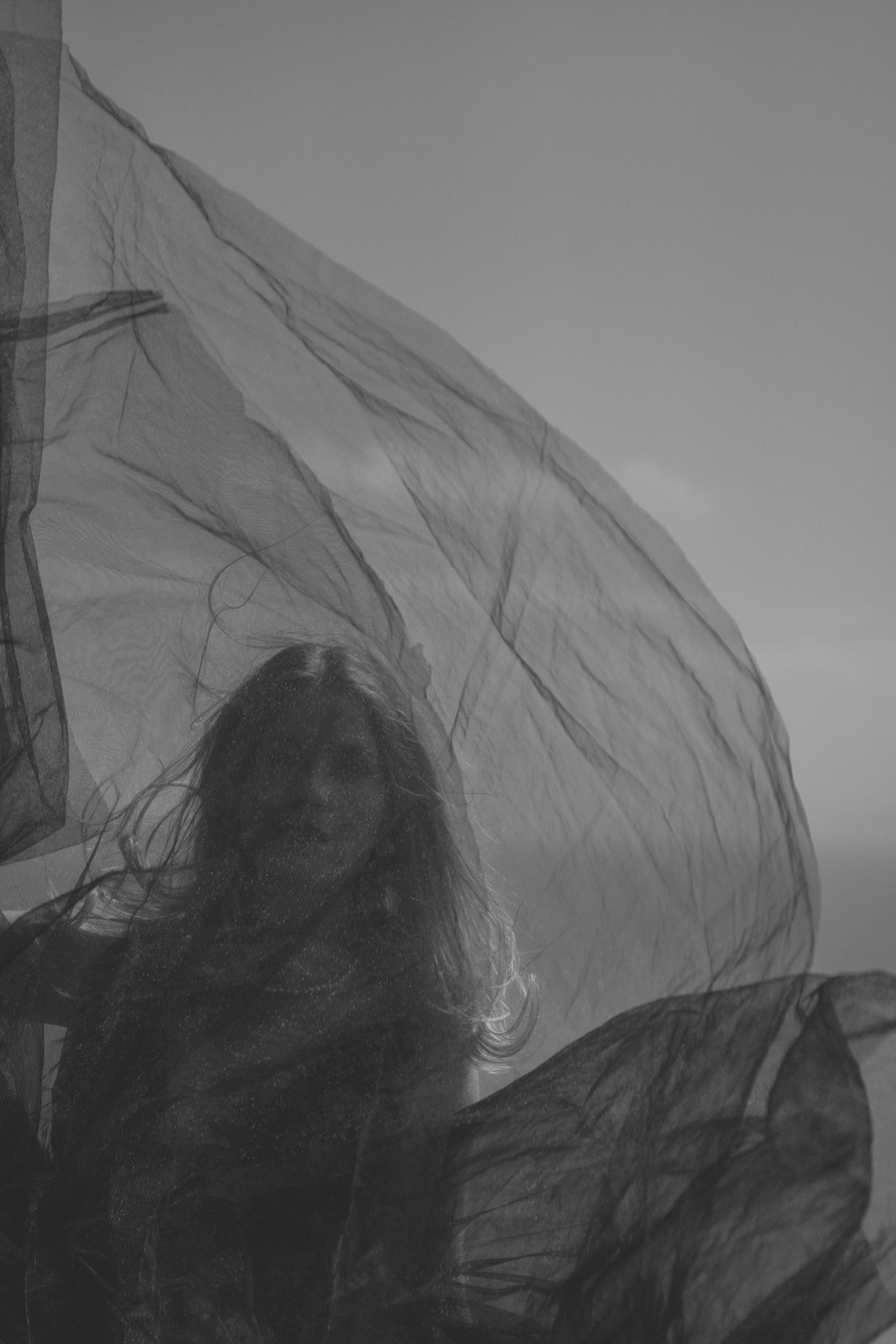 A silhouetted figure stands behind flowing sheer fabric in a moody black and white artistic portrait.
