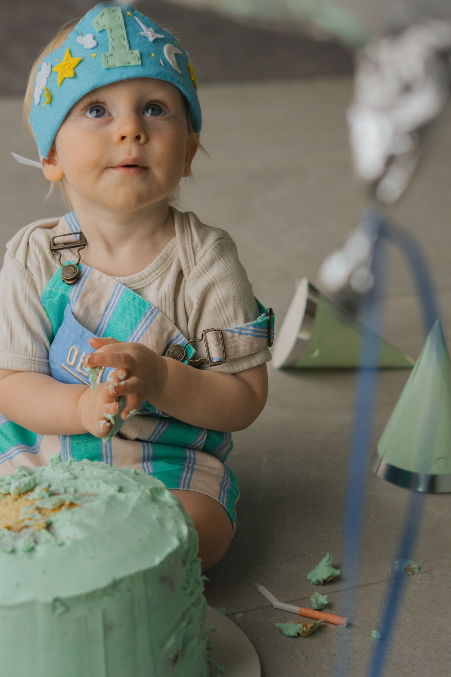 Baby in blue star hat enjoys first birthday cake smash with mint green frosting and blue decorations.