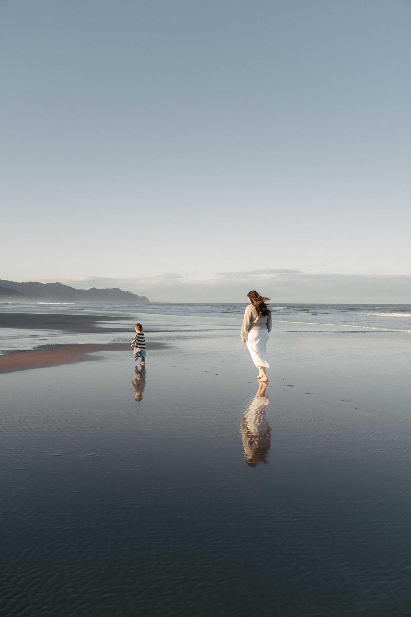 A woman and child walk on a reflective wet beach at sunset with misty mountains in the background.