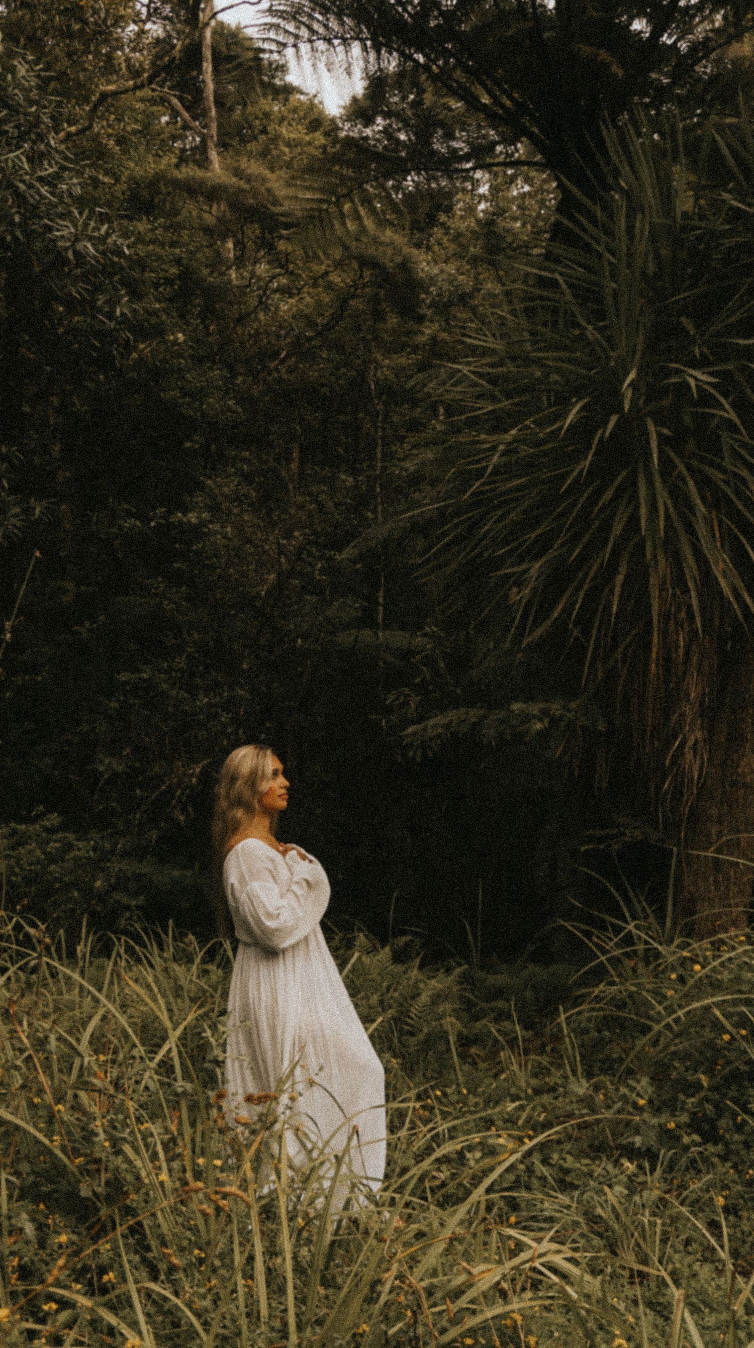 Figure in white dress stands among wild grasses at sunset with palm trees in background.