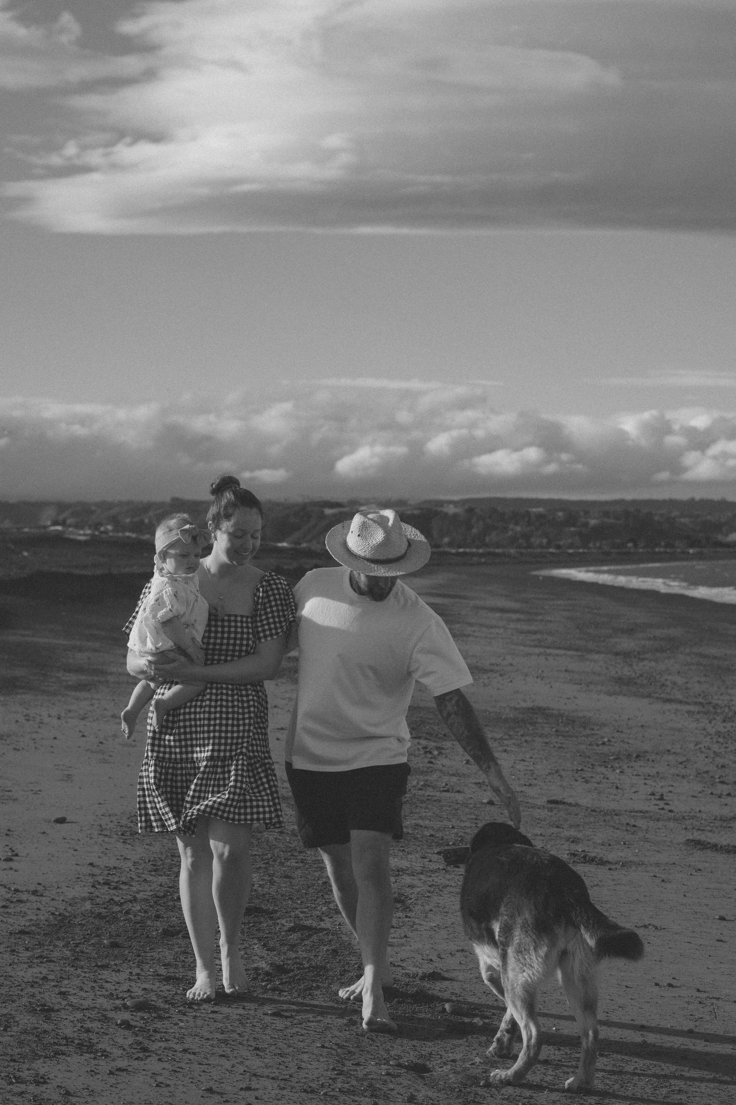 A black and white sequence of photos showing two people walking with a dog along a beach at sunset.