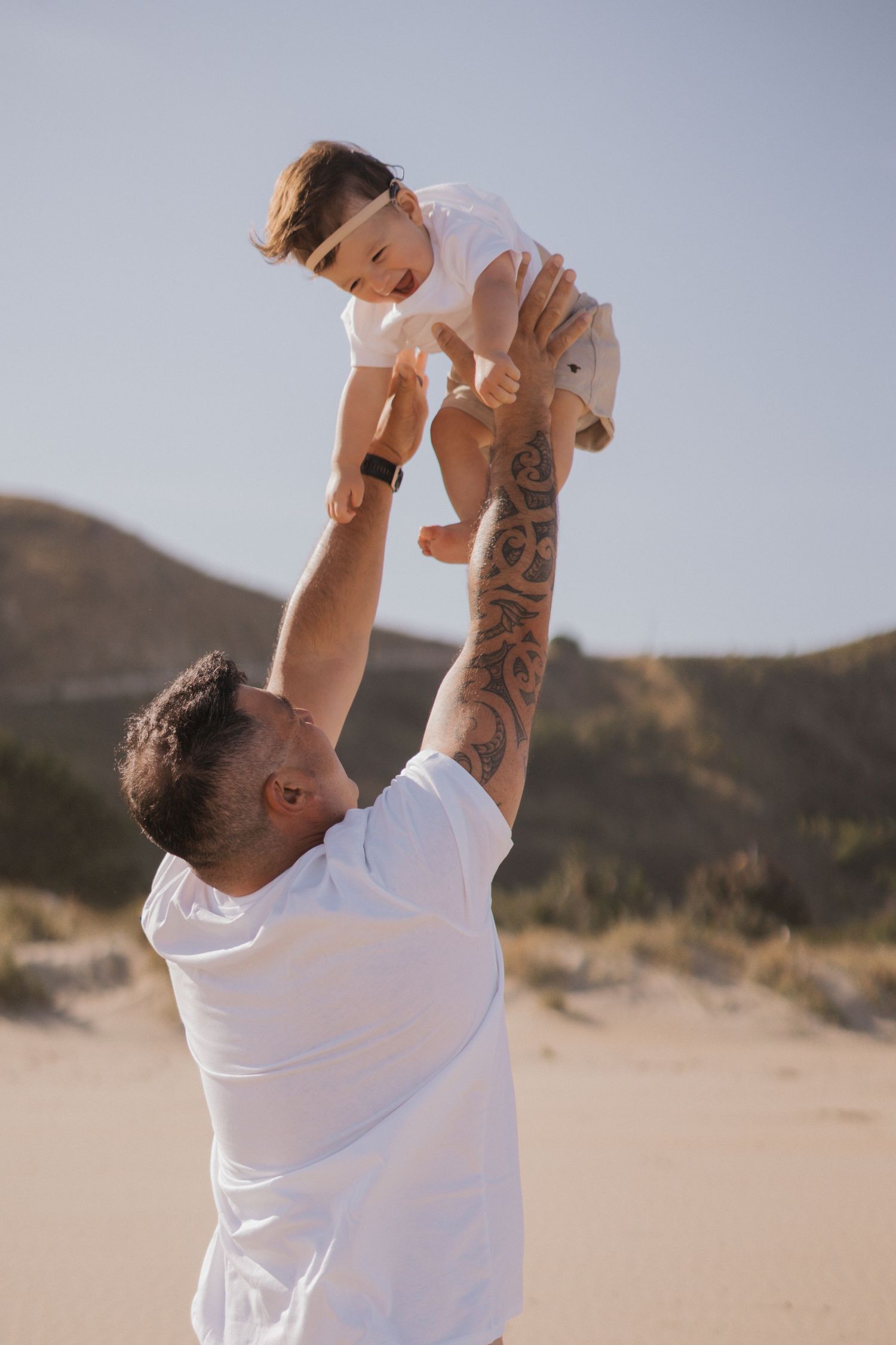 A person in a white shirt lifts a small child into the air against a desert landscape at sunset.