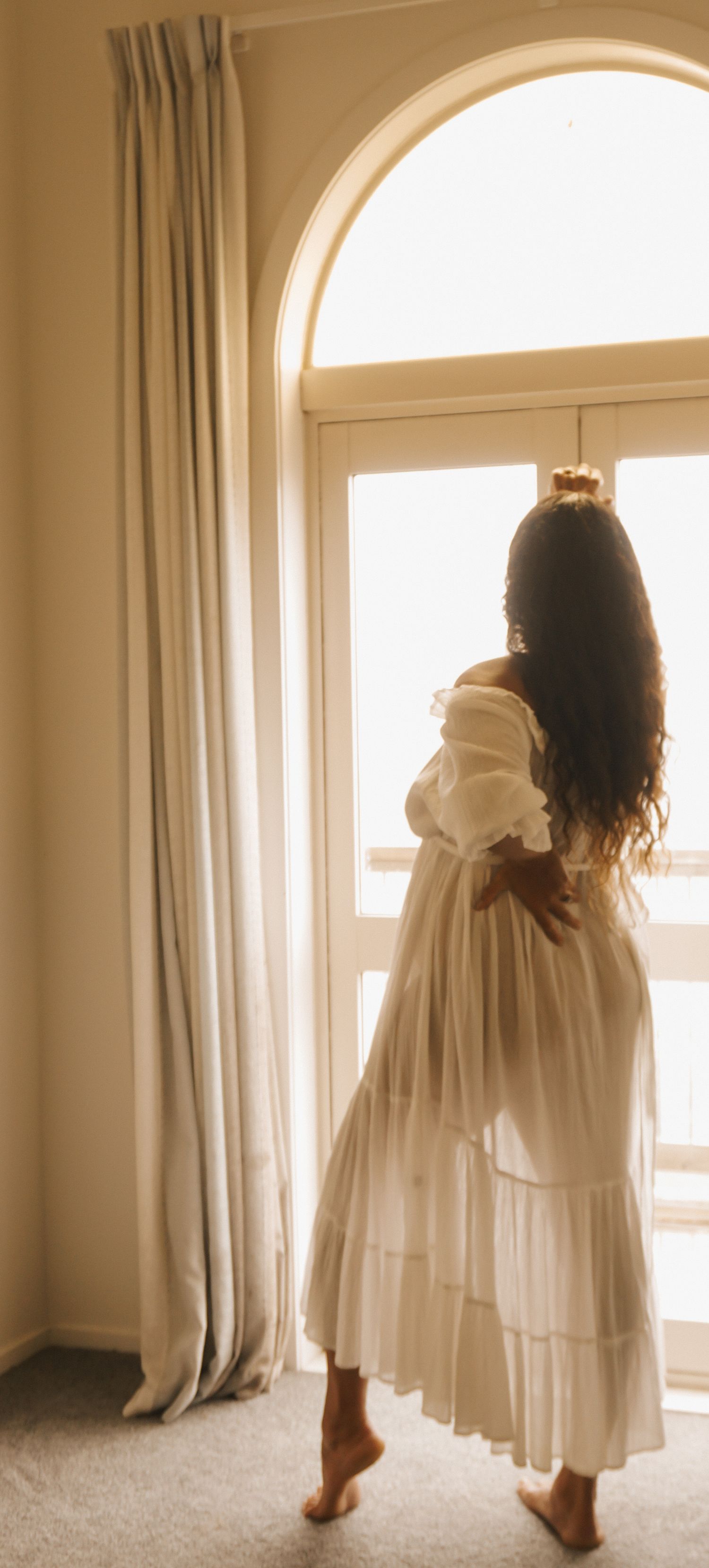 Artistic maternity photos in white dress by arched windows with soft lighting.