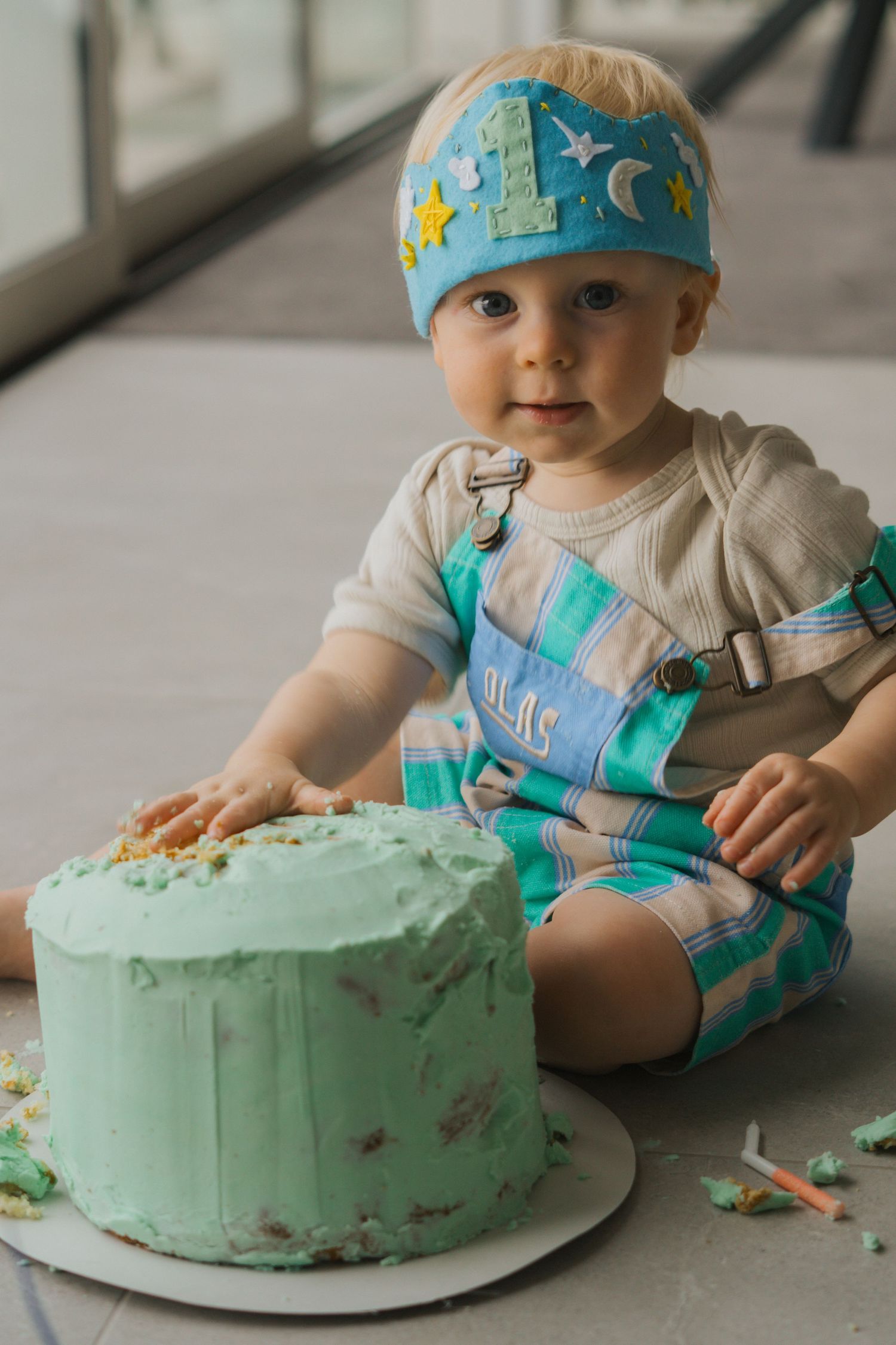 A baby in a blue birthday hat sits on the floor with a mint green frosted cake, showing a cake smash photo sequence.