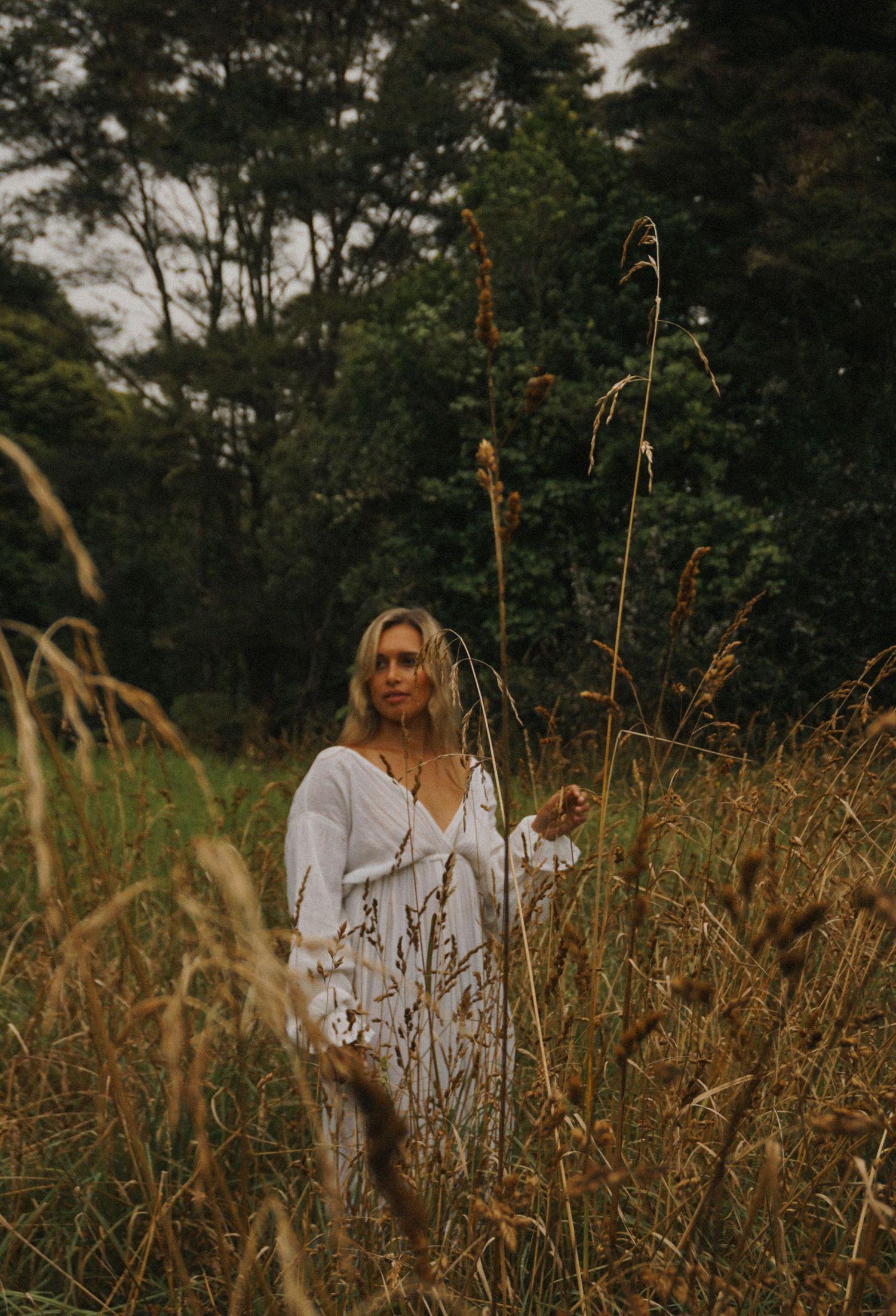 Person in white dress poses amid golden wild grass during sunset.