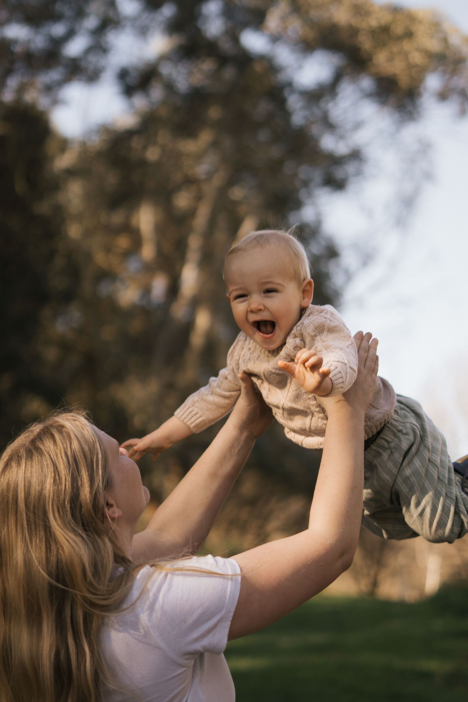Parent lifting and playing with a laughing baby outdoors in golden sunlight.