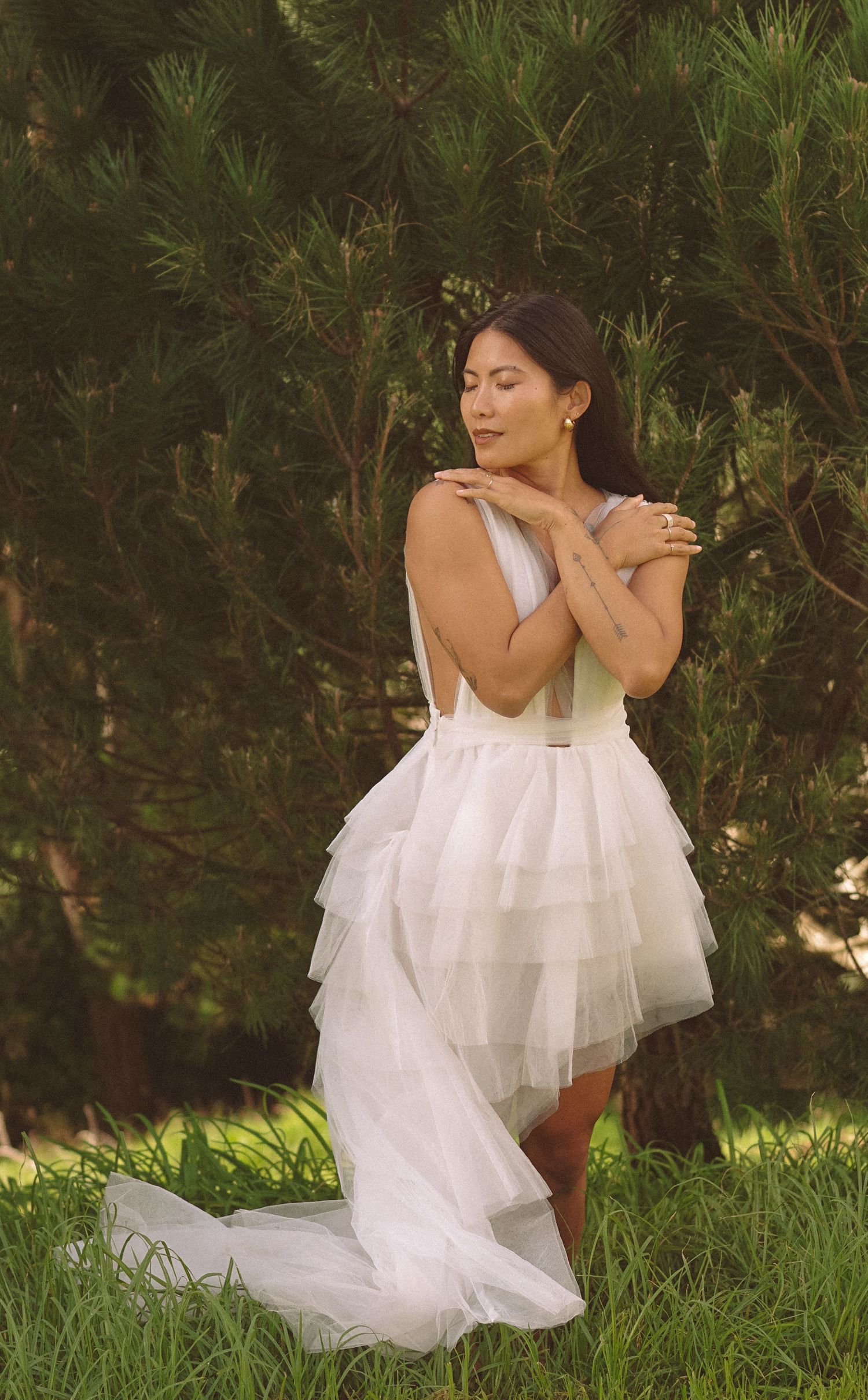 Figure in flowing white dress sits gracefully on grass surrounded by greenery.