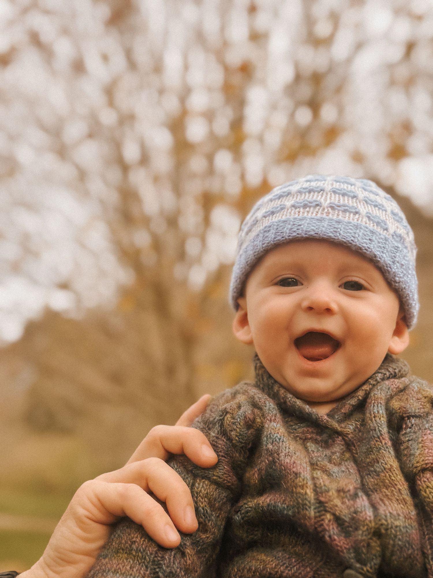 A baby wearing a knit hat and sweater smiles and laughs outdoors.