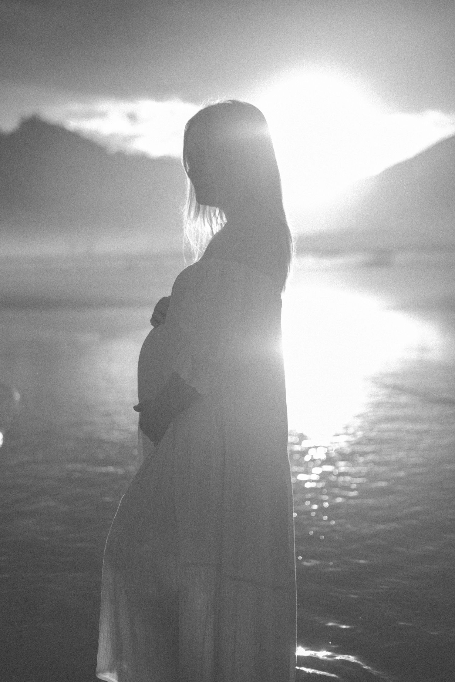 A silhouetted pregnant figure stands against a bright sunset over water in a dreamy black and white photograph.
