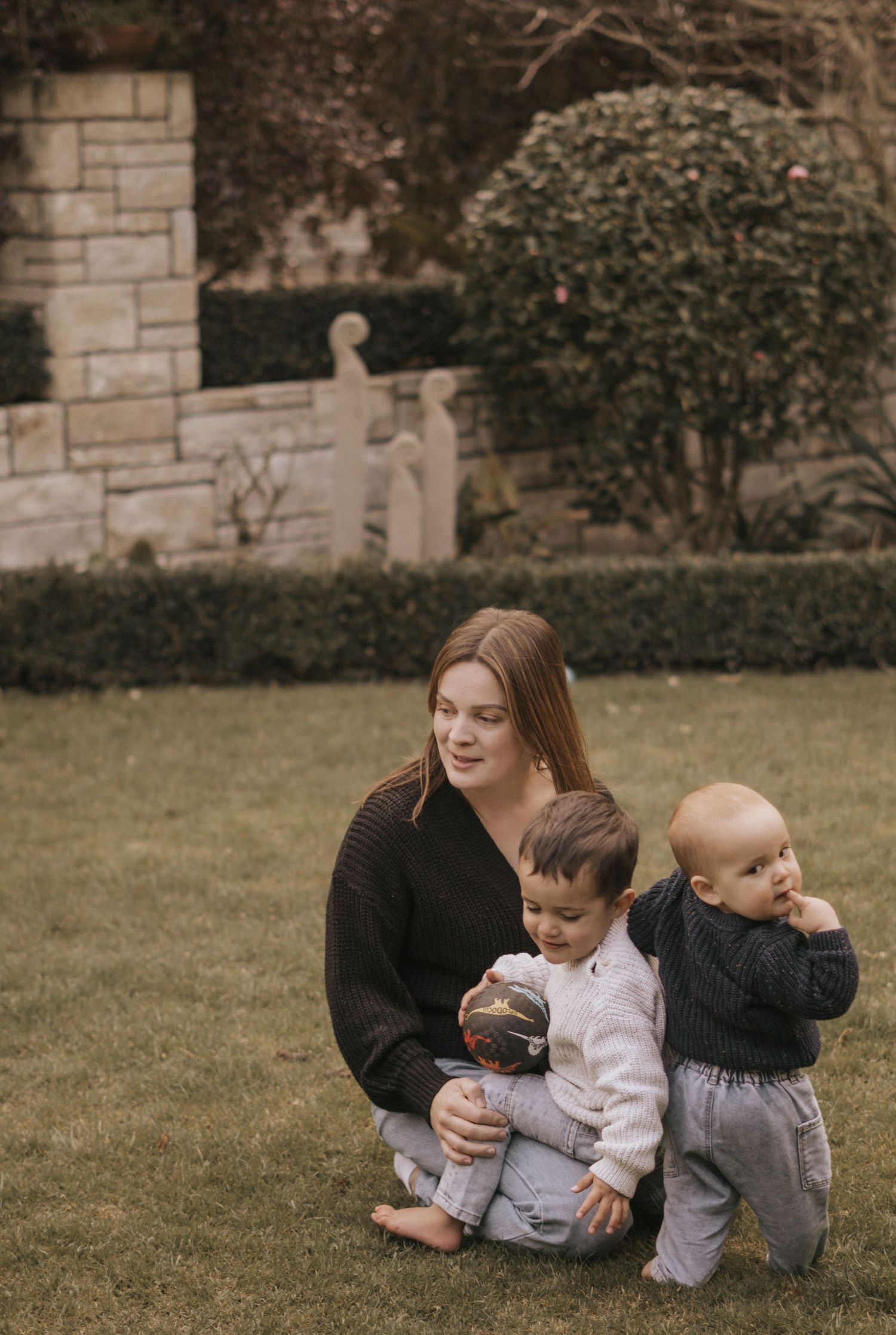 Family playing and laughing together on grass near a stone wall outdoors.