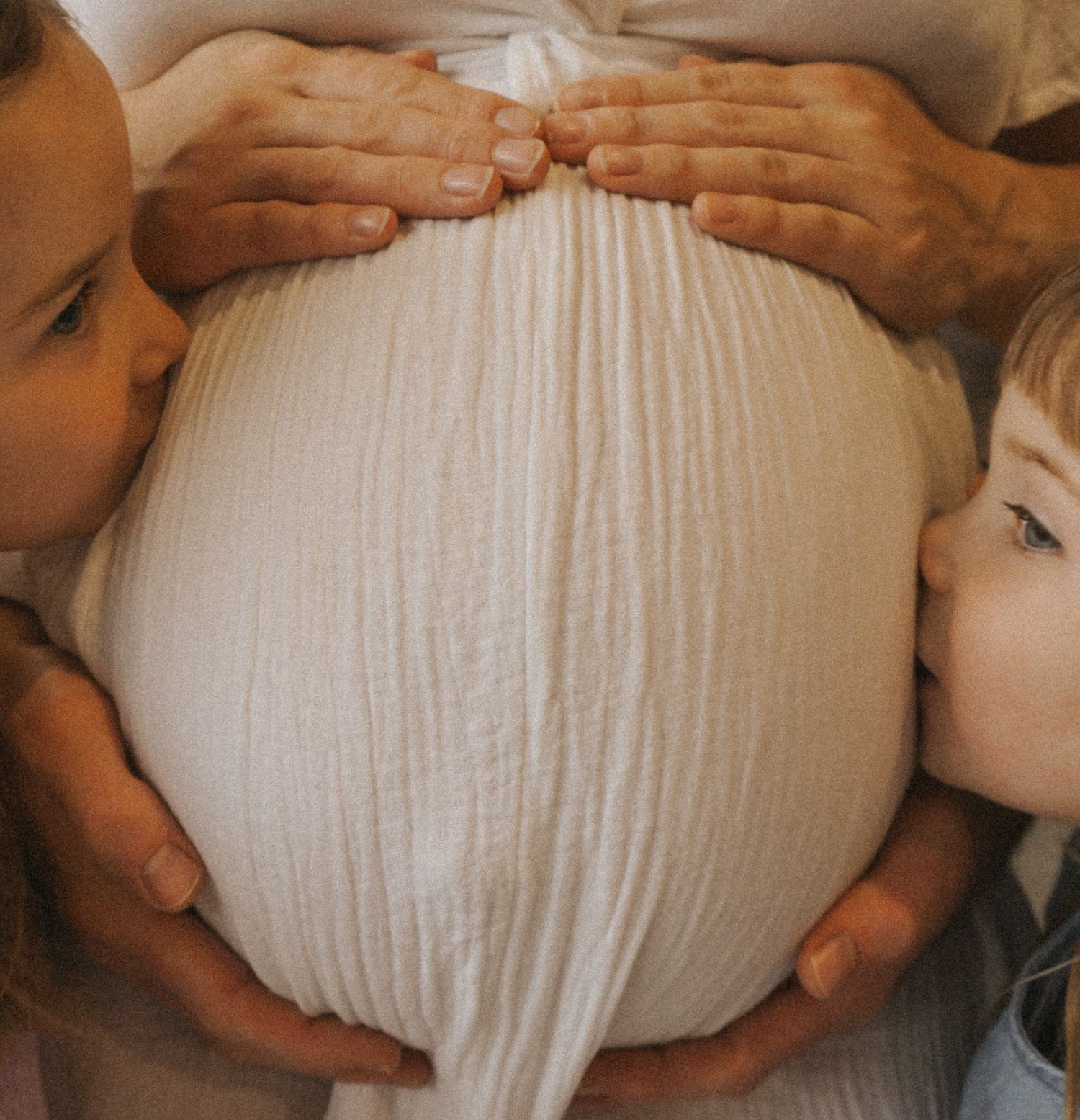 Multiple hands gently cradling a pregnant belly in white dress.