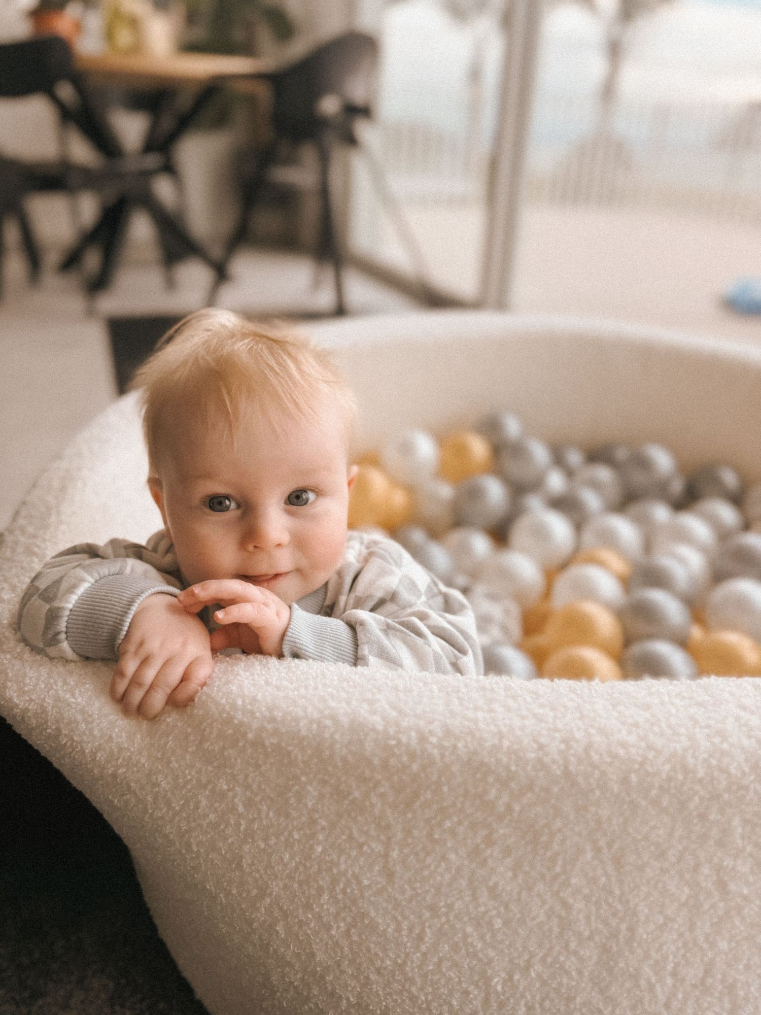 A baby peeks over the edge of a white ball pit filled with neutral colored balls.