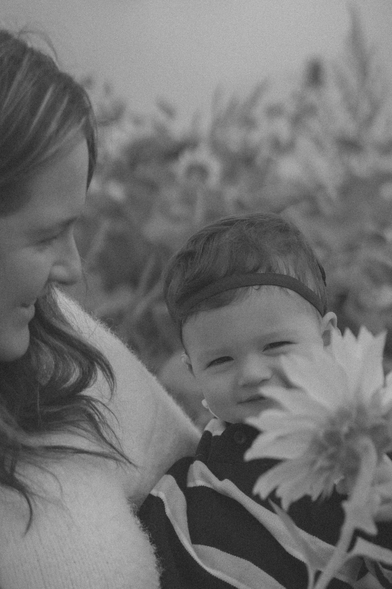 A tender black and white portrait of a parent and baby among sunflowers in a garden setting.