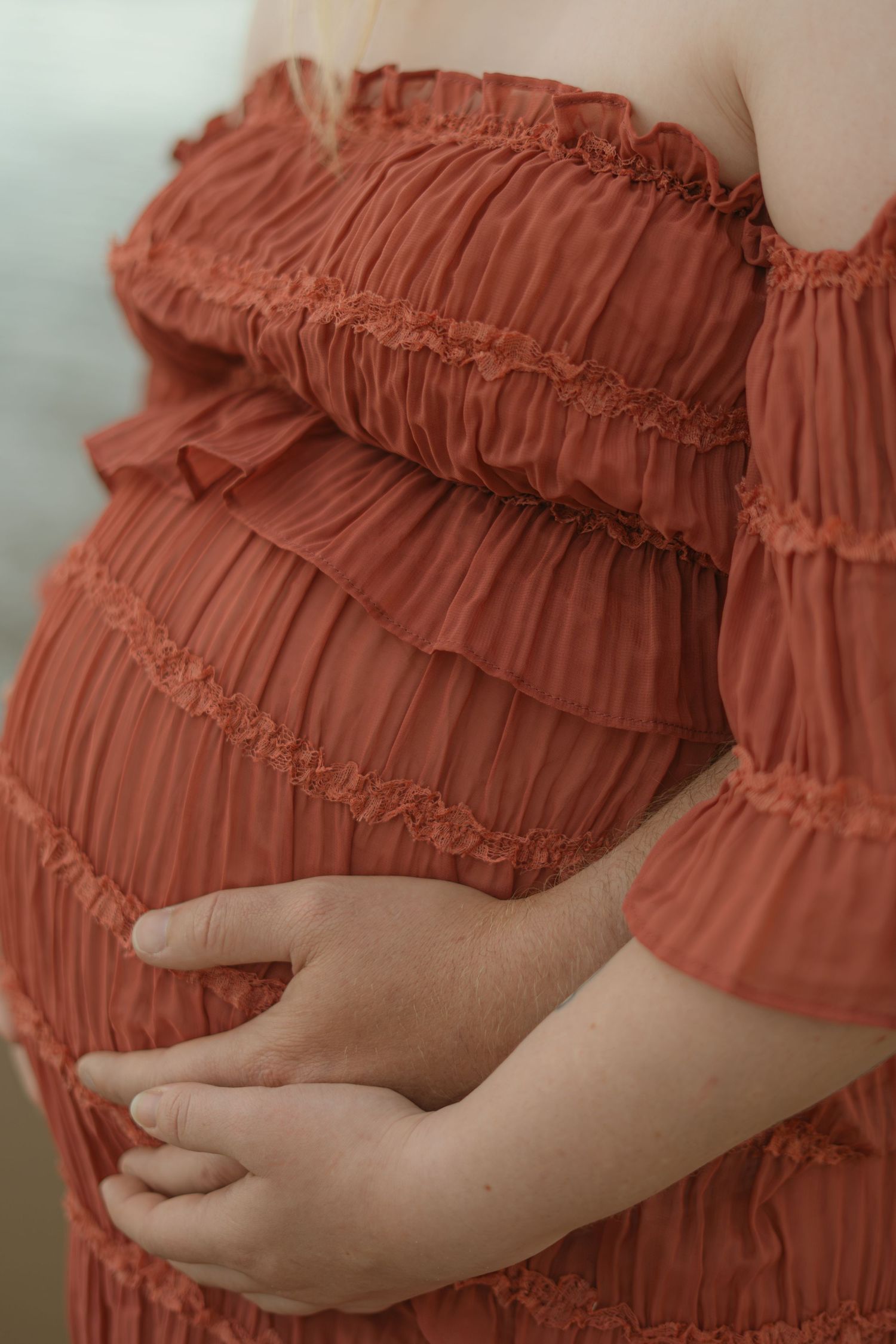 Close-up of pregnant belly covered in coral-colored ruched fabric dress with hands gently cradling the bump.