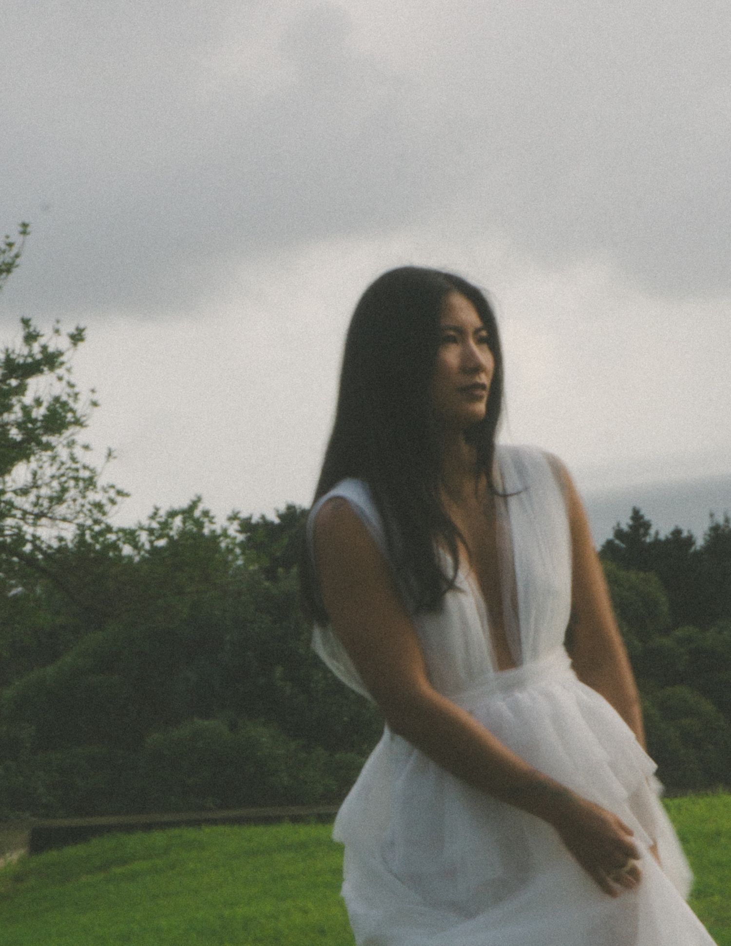 Figure in flowing white dress twirls joyfully in a green field under cloudy skies.