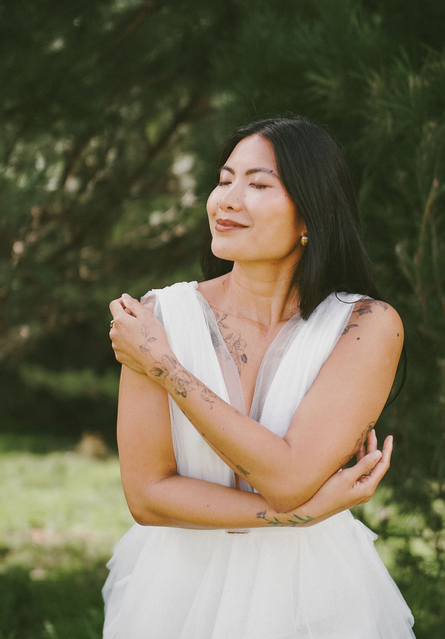 Person in white dress embracing themselves in a sunlit garden setting.