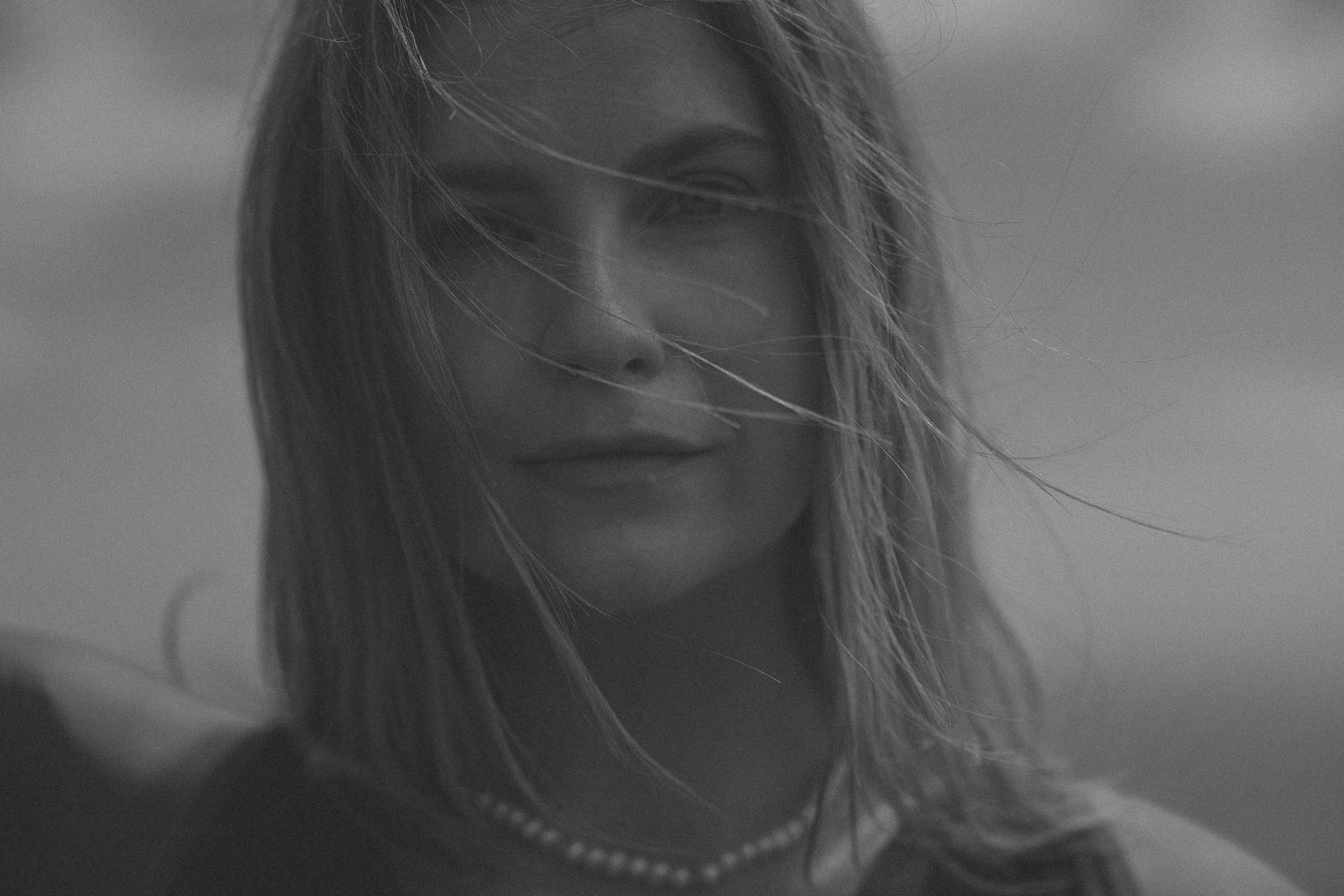 A black and white portrait of someone with windswept hair wearing a necklace, shot with soft focus.
