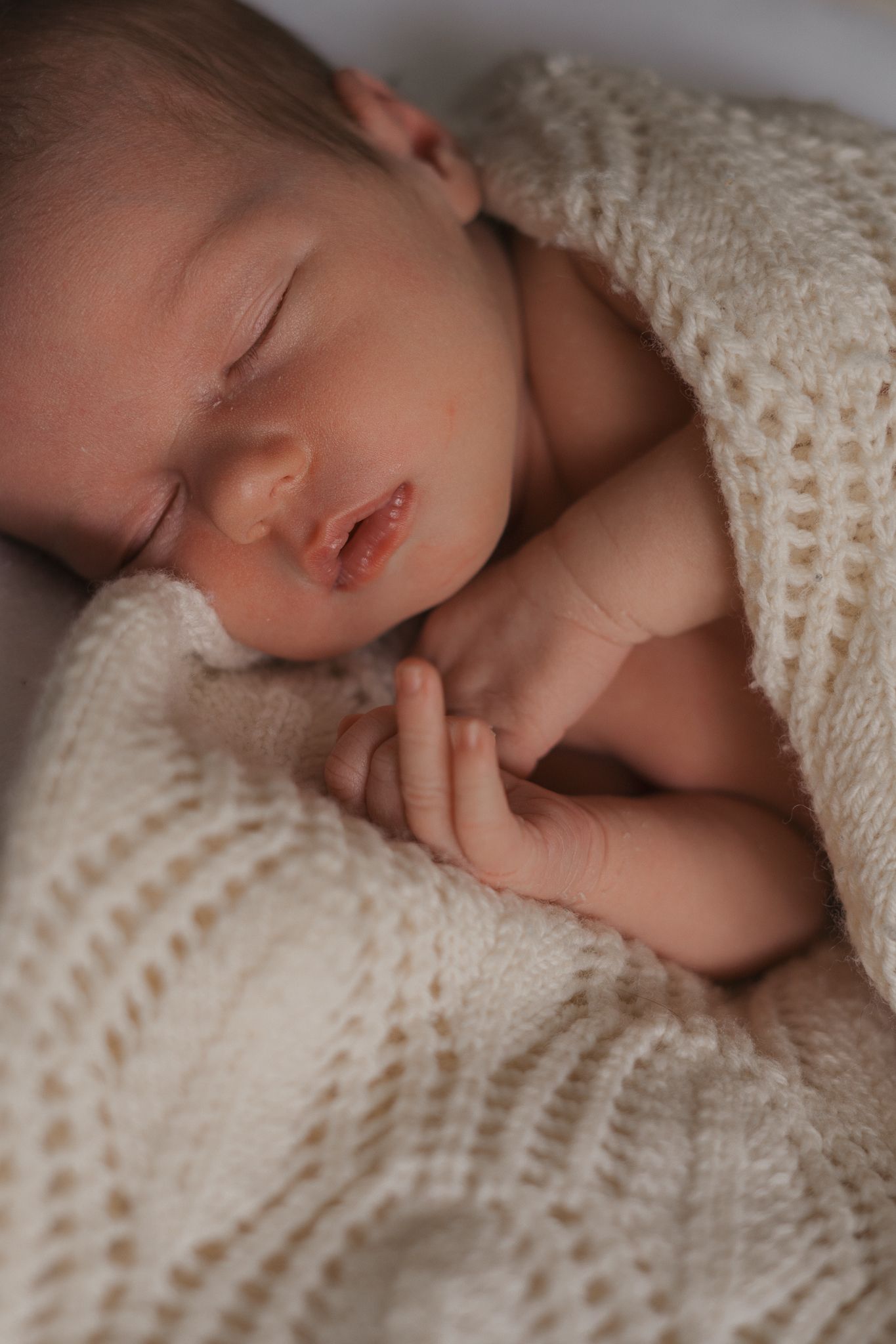 A sleeping baby wrapped in a cozy cream-colored knit blanket rests peacefully in soft natural light.