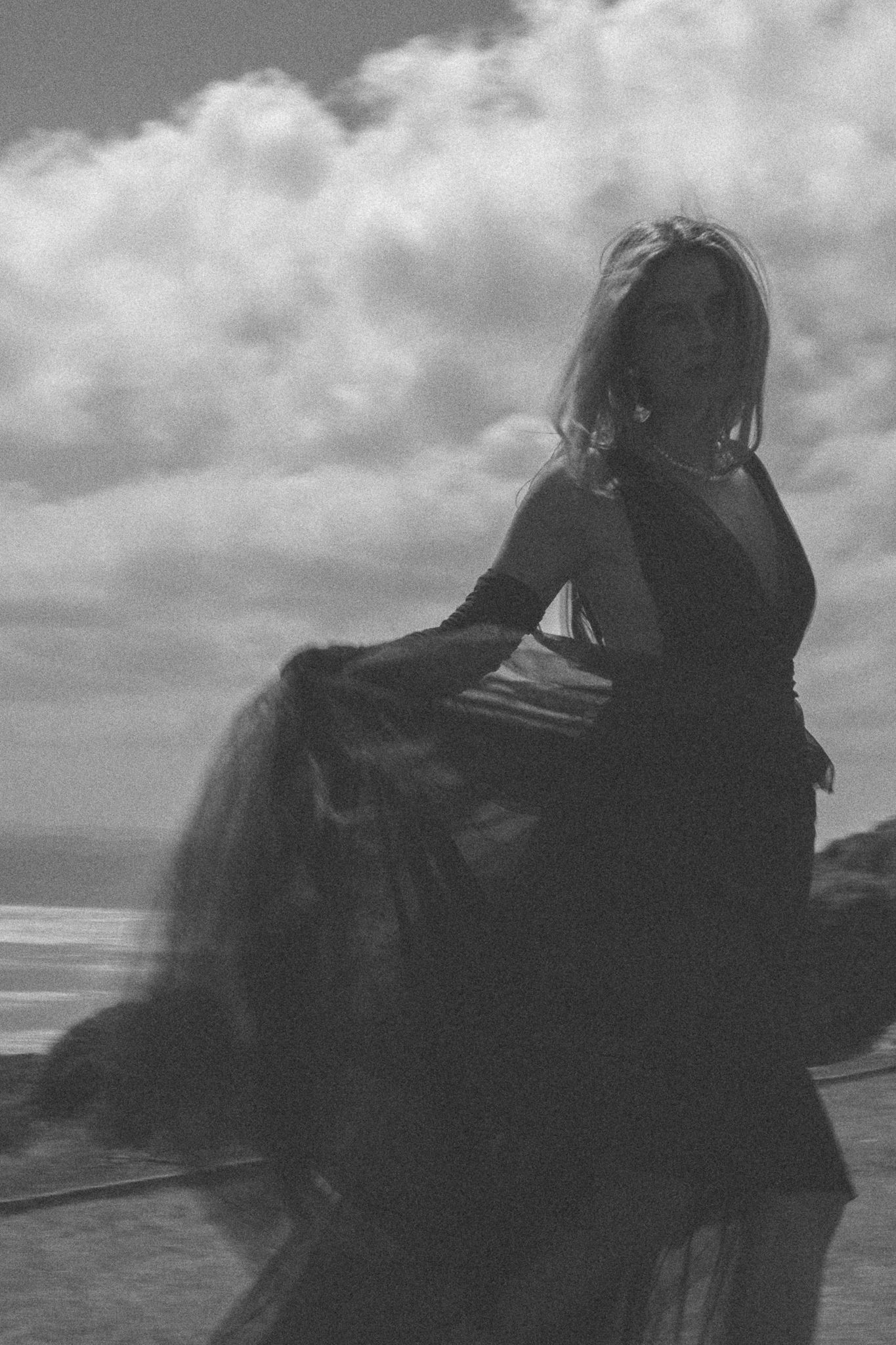 A silhouetted figure in a flowing dress poses dramatically against a cloudy sky by the ocean.
