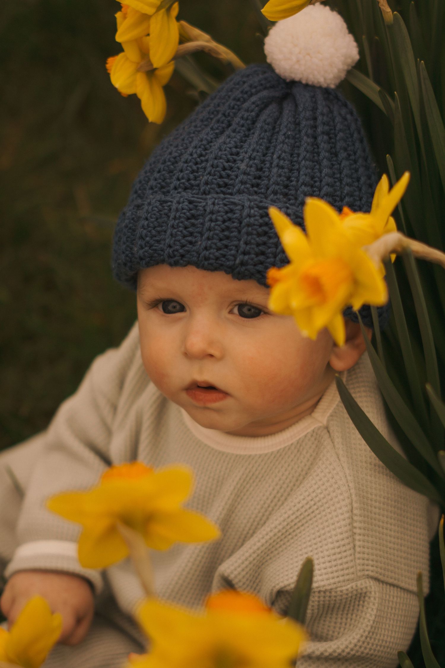 Baby wearing blue knit hat among bright yellow daffodils.