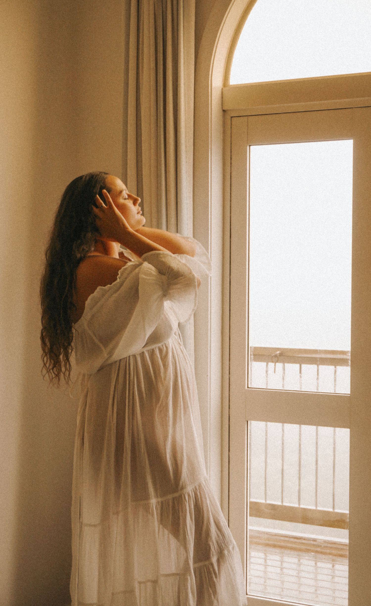 Woman in white dress basking in sunlight by window in serene moment.