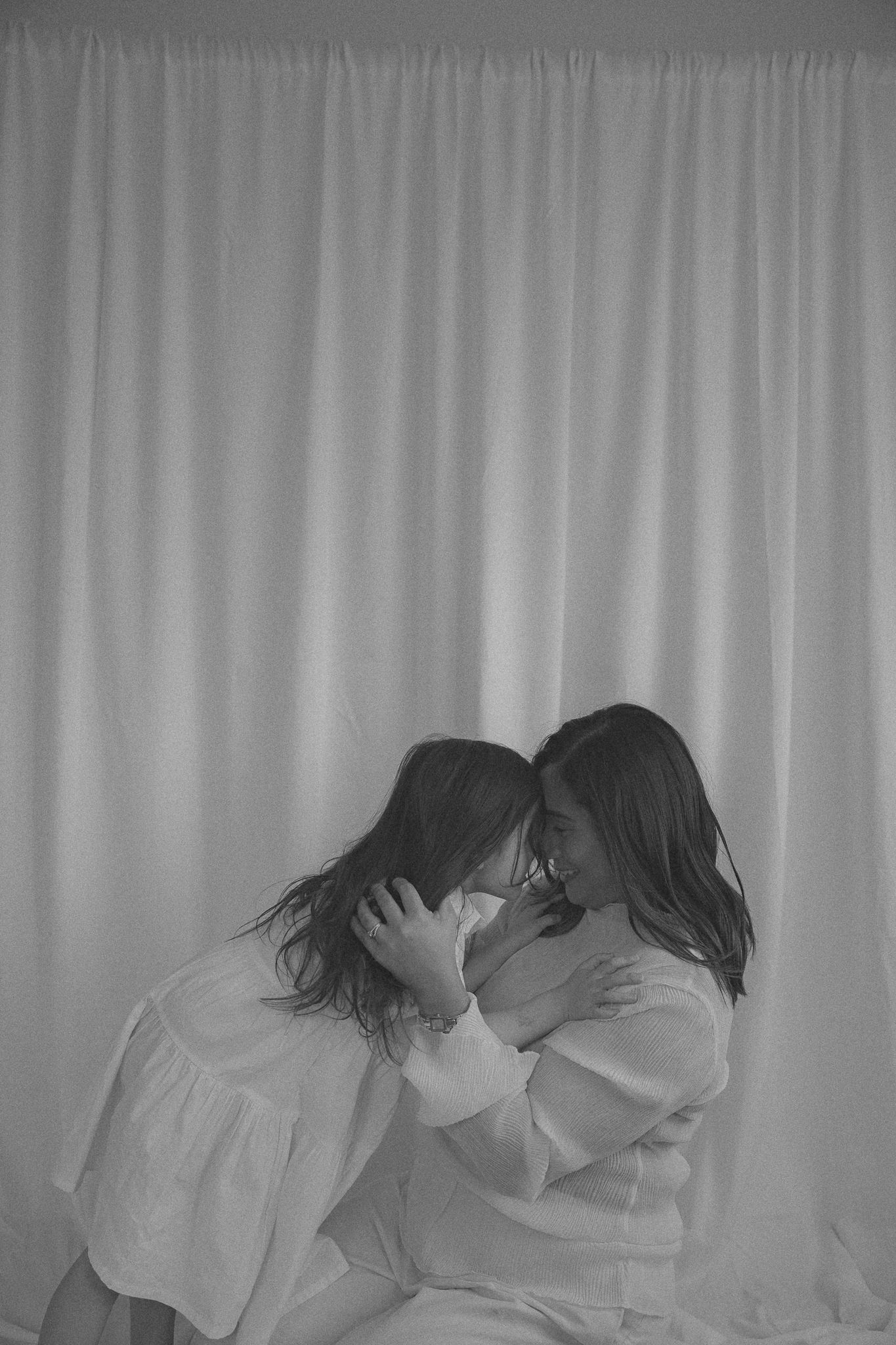 Black and white photo of two young girls in white dresses embracing tenderly against a soft curtain backdrop.